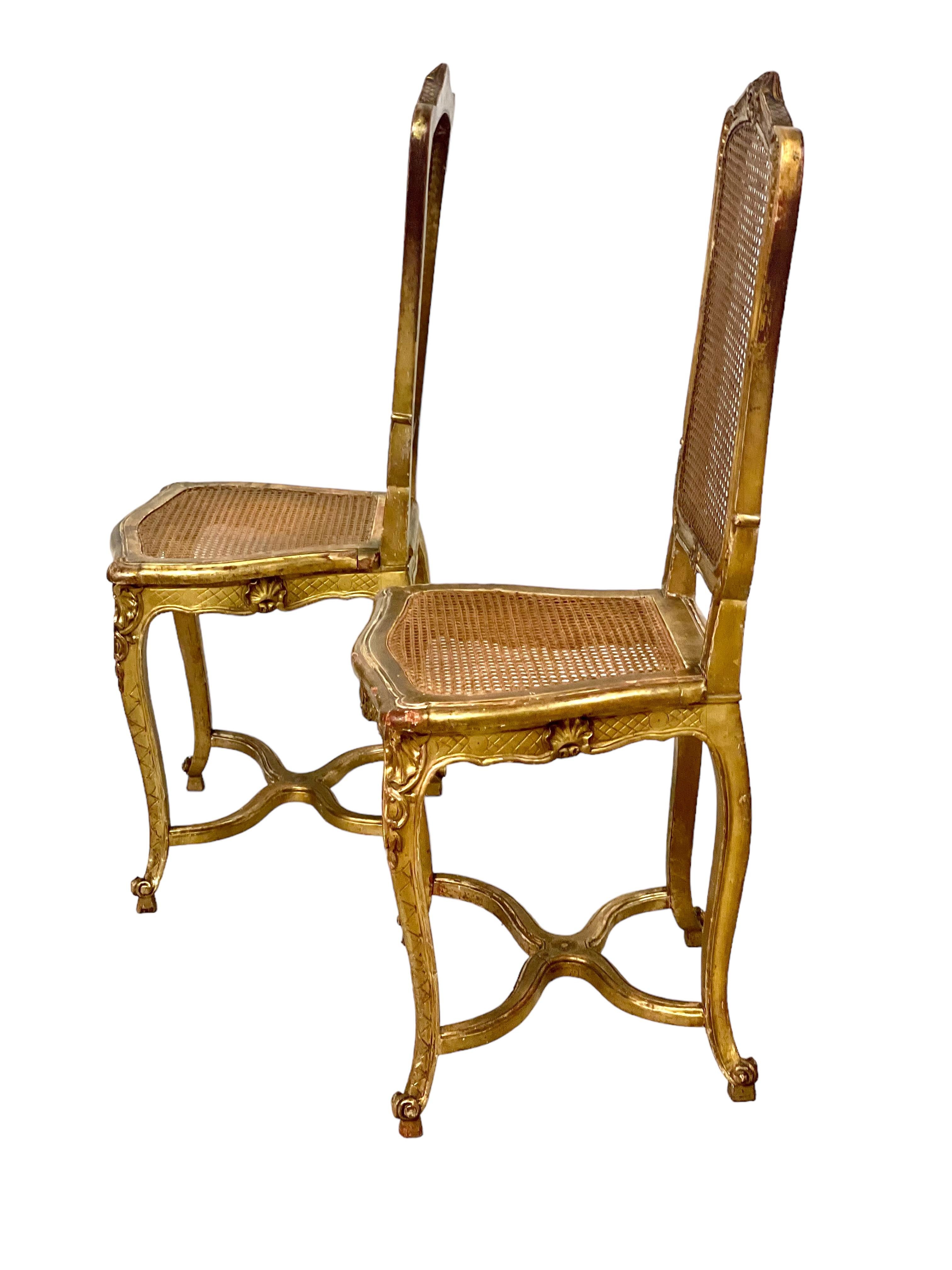 A delightful pair of late 19th century carved and moulded giltwood side chairs, with caned seats and backrests. These elegant chairs, with their high, cartouche-style backs, are raised on cabriole legs joined by a delicately curved X-shaped
