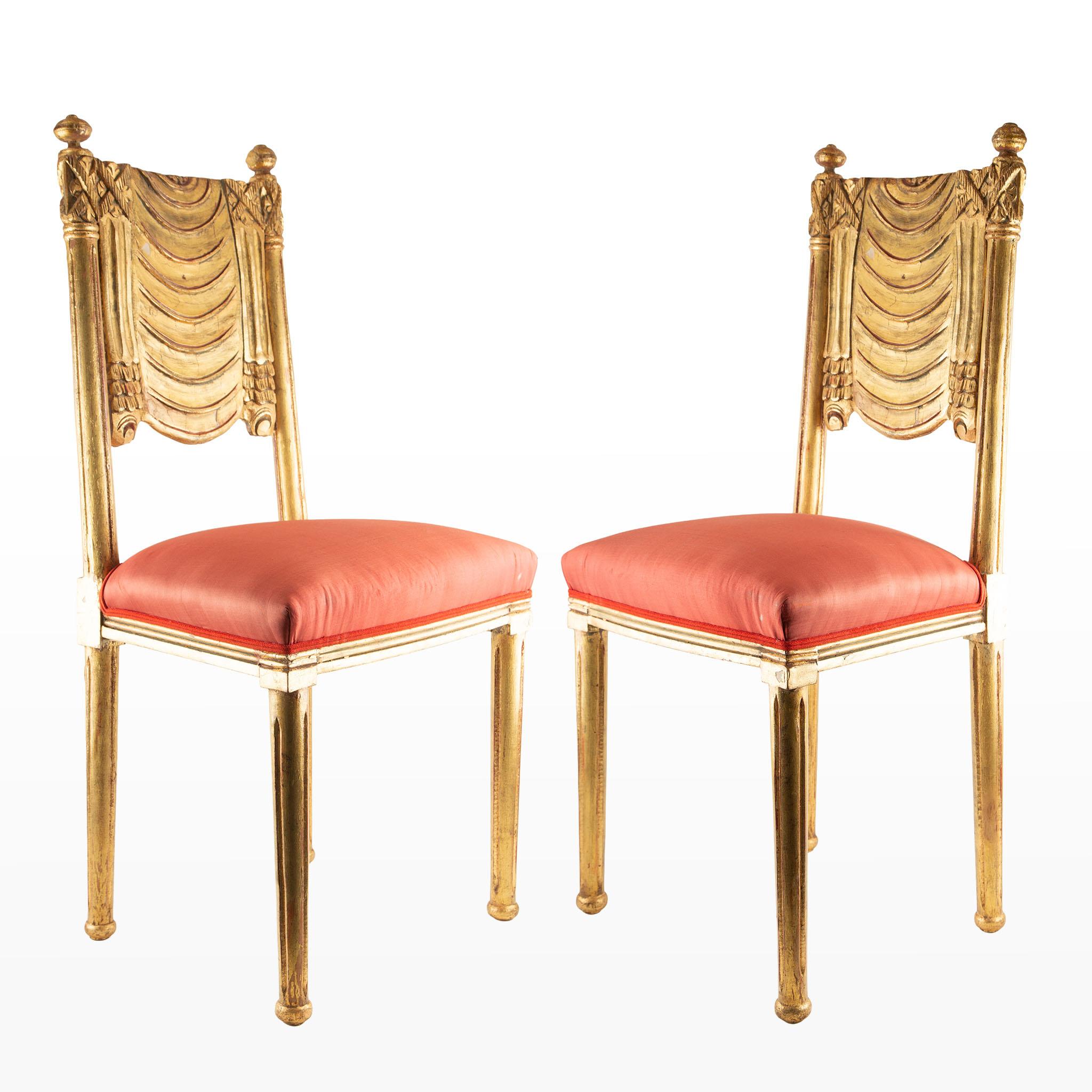 Pair of French Art Deco giltwood side chairs, circa 1930s, featuring elegantly carved backs with stylized draped swag motifs and finial-topped stiles, all finished in a rich gold leaf. The seats are upholstered in vibrant red silk, adding a bold