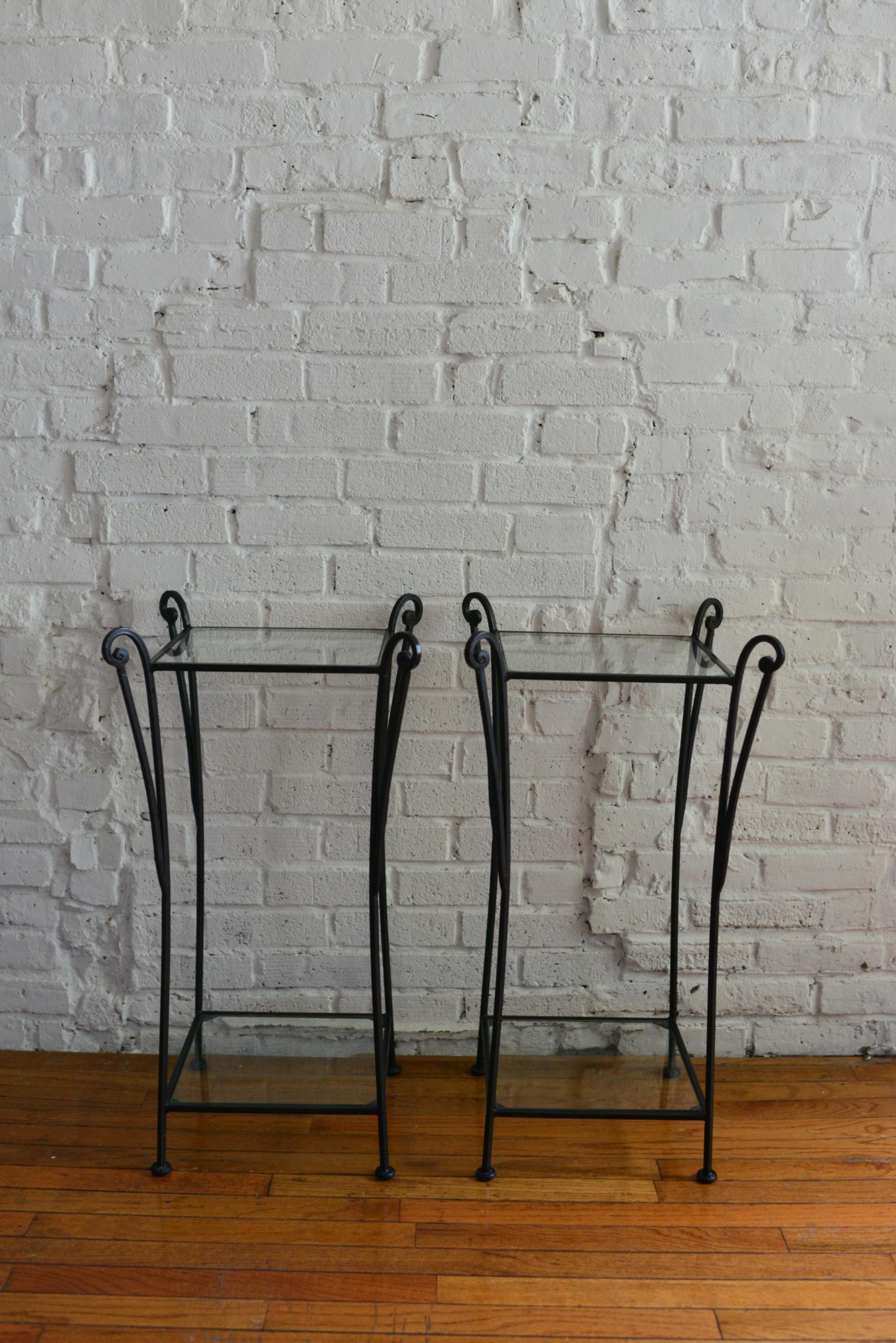 Unique and playful pair of wrought iron and glass pedestal side tables. The two-tier tables feature a wrought iron base with dramatically curved legs and curve details. Each table has an inset glass tabletop and shelf. Similar in style to many