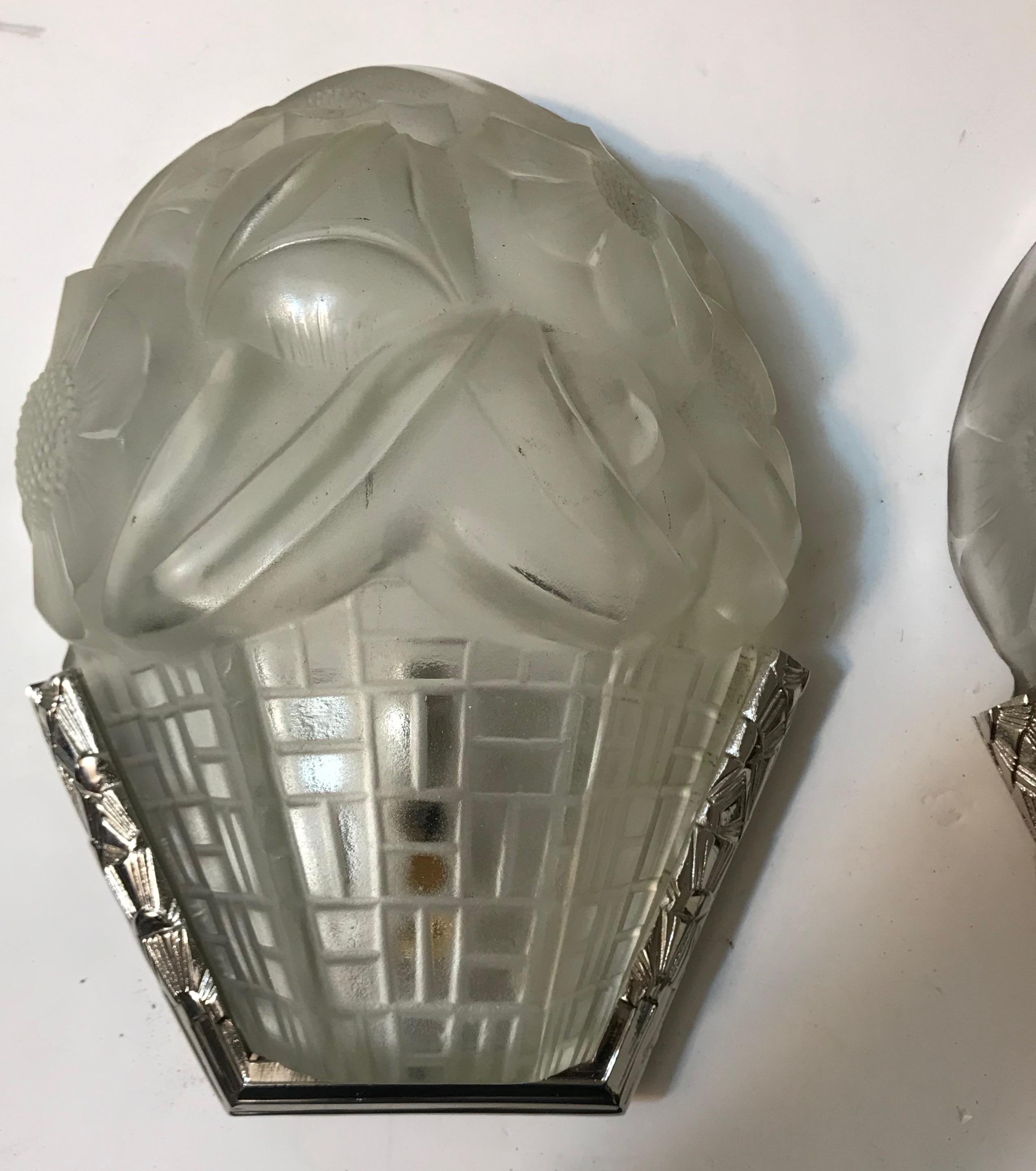 Pair of French Art Deco wall sconces signed by the French artist “Degue” in clear frosted glass shades with floral motif. Held by Polished nickel design frames. Has been rewired for American use with one candelabra socket. Each socket has max watt