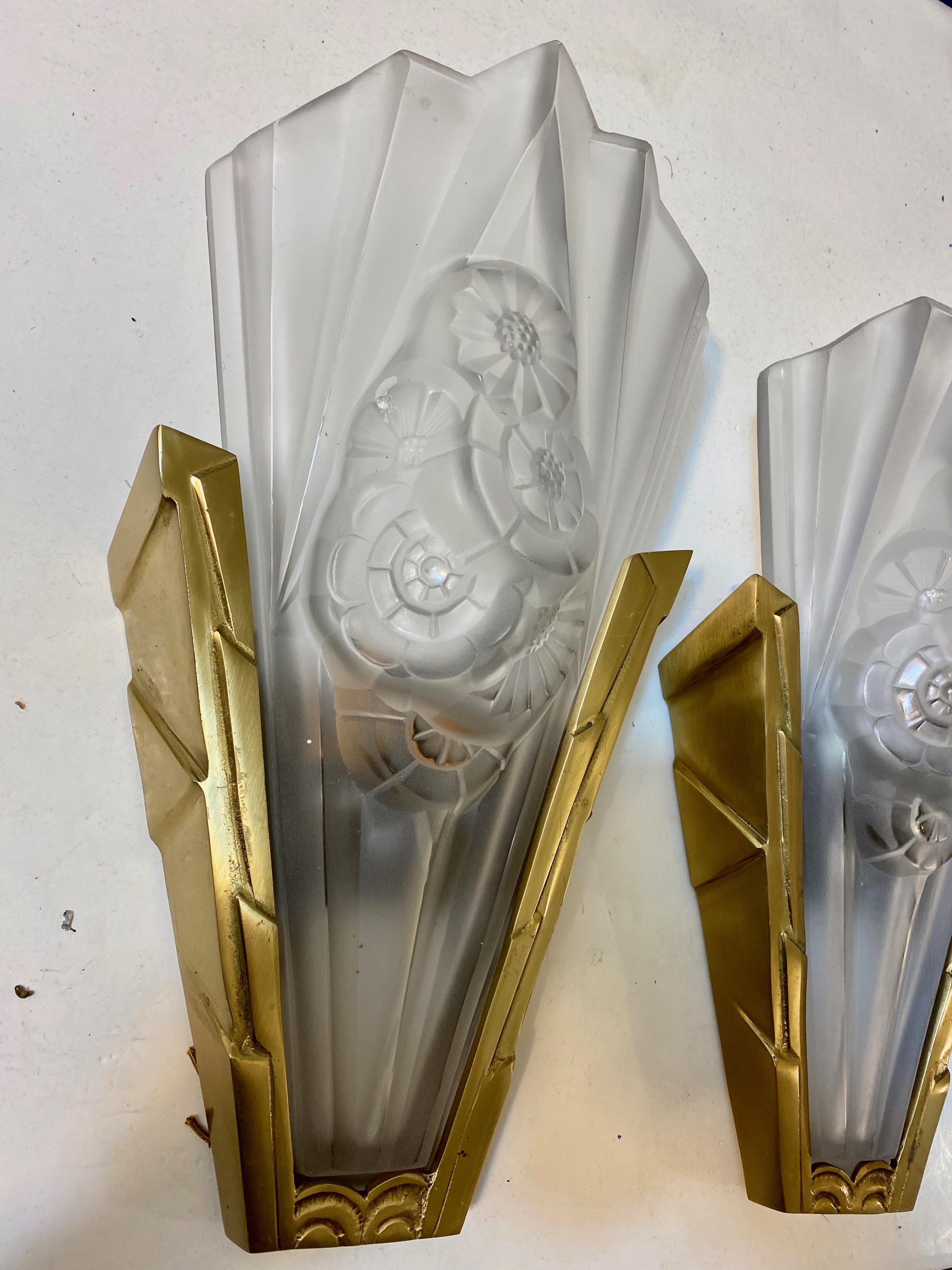 Pair of French Art Deco wall sconces by the French artist Degue. Clear frosted glass shades decorated with geometric and floral motifs. Held by geometric brass design frames. Each glass shade is marked DEGUE. Has been rewired for American use with