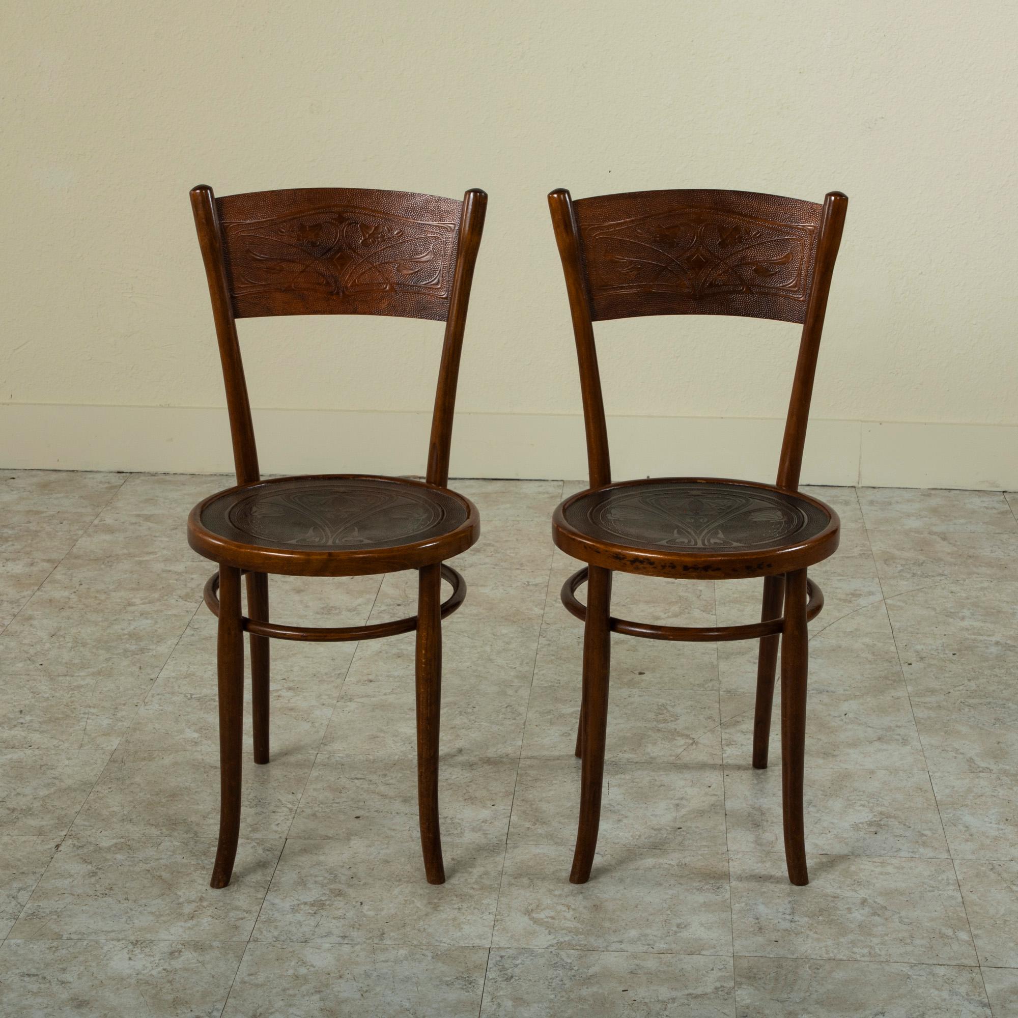 This pair of French Art Nouveau period bentwood bistro chairs features pressed wood seats and seat backs detailed with classic stylized lilies and scrolling leaves. The seats rest on gently curved legs joined by a circular support. The chairs bear