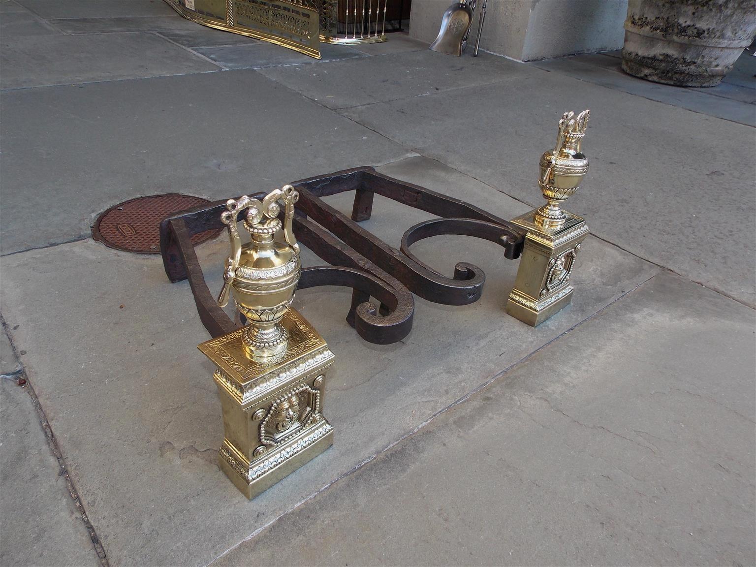 Pair of French brass and wrought iron andirons with flanking ribbon and foliage urns, hand chased floral beading, squared plinths with figural lions head, circular corner medallions, and resting on step back floral base. Andirons retain the original