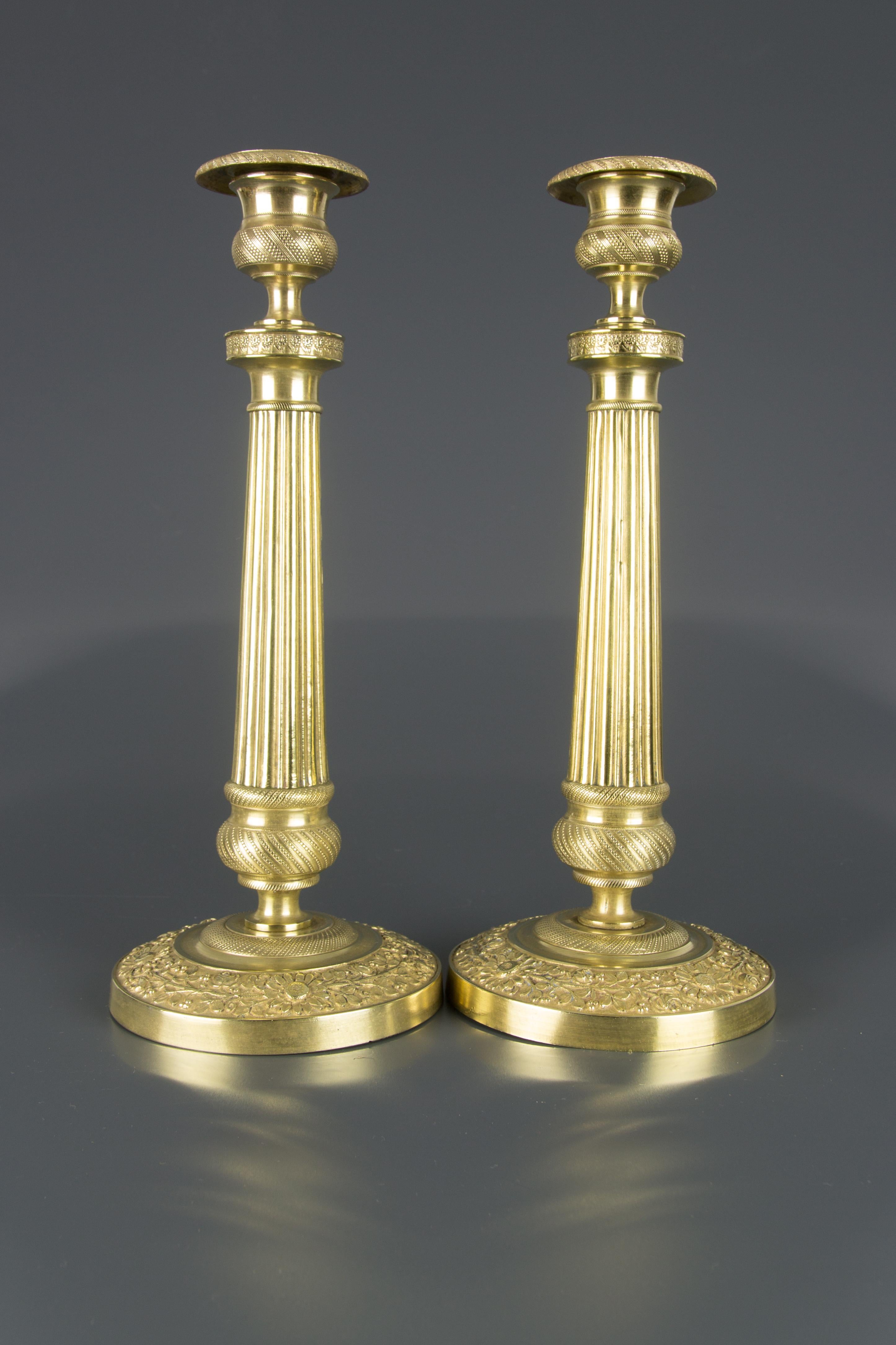 A beautiful pair of French Restauration style brass candlesticks with removable bobeches (drip trays). Each candlestick features a fluted columnar stem, has an ornate collar, decorated with flowers, and is resting on a circular base highlighted with