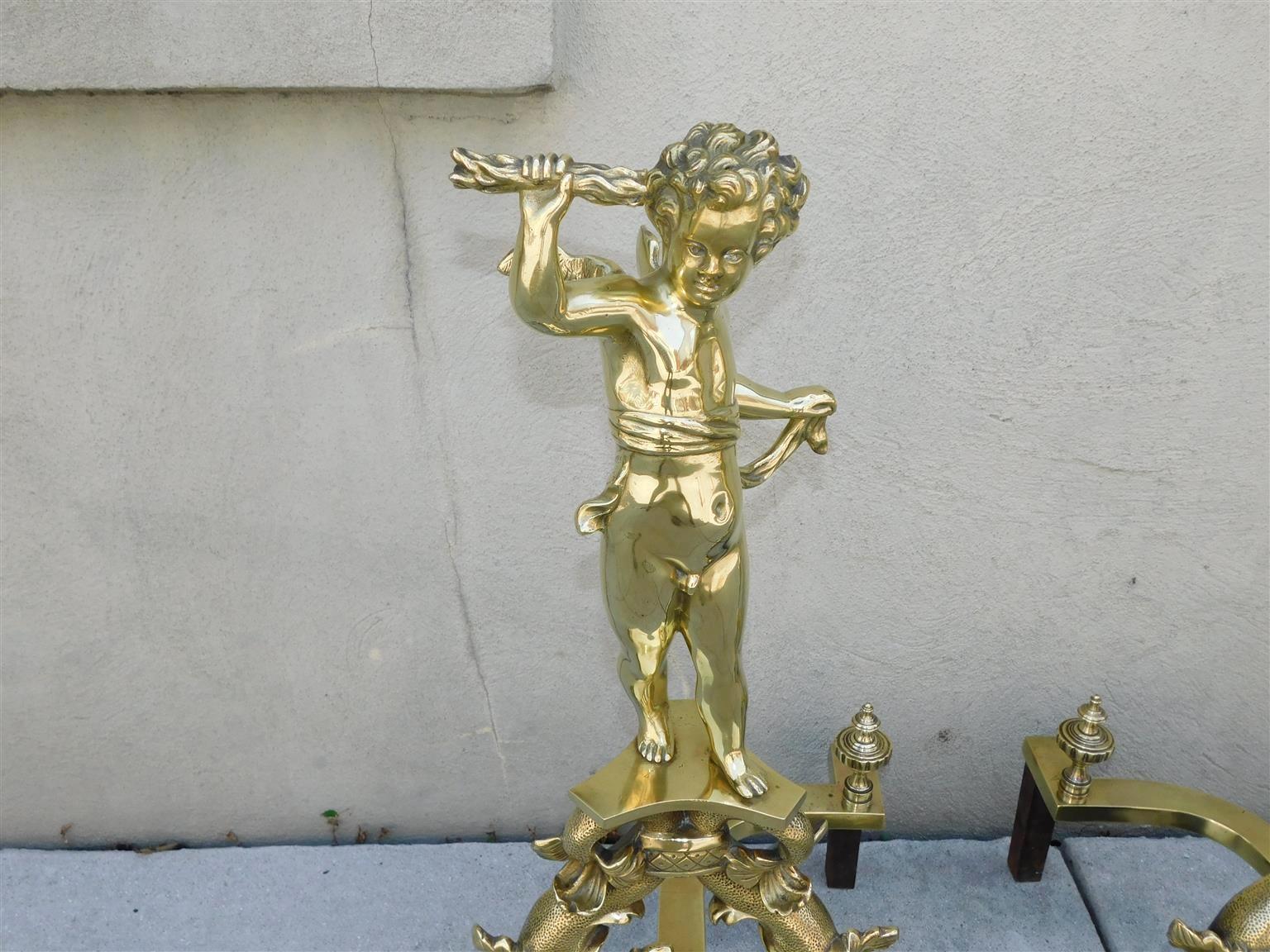Pair of French brass decorative andirons with flanking cherubs holding scrolls, intertwined banded dolphins, and resting on the original curvature brass and iron rear legs with matching urn fluted log stops, Early 19th Century