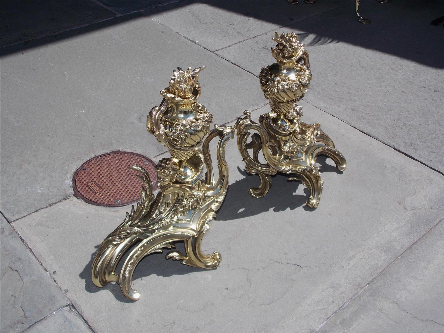 Pair of French brass flame urn finial chenets with flanking winged cherubs and decorative scrolled floral motif, Early 19th century.