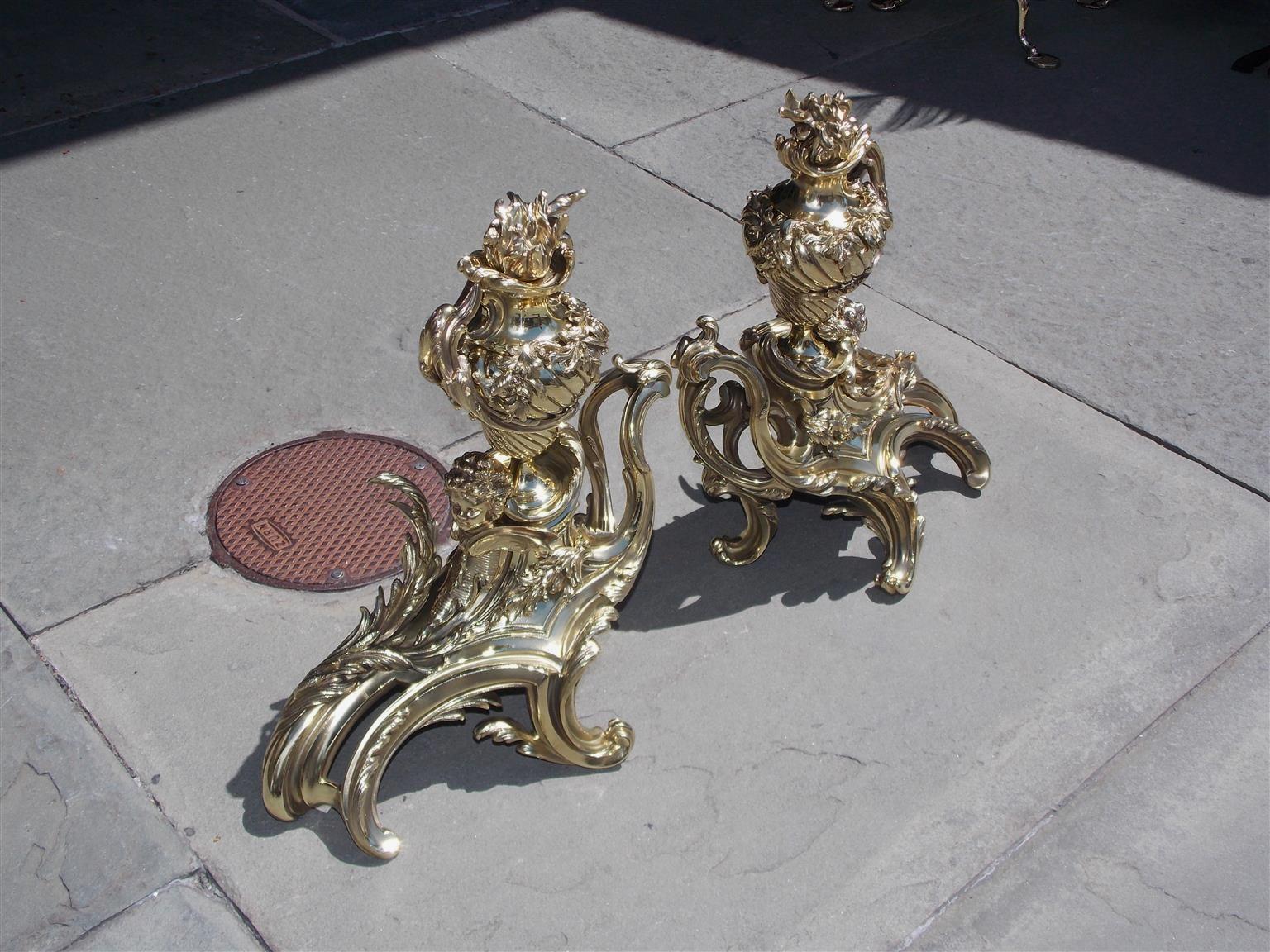Coppia di chenet a forma di urna a fiamma in ottone con cherubini alati affiancati e motivo decorativo floreale a volute, inizio XIX secolo.