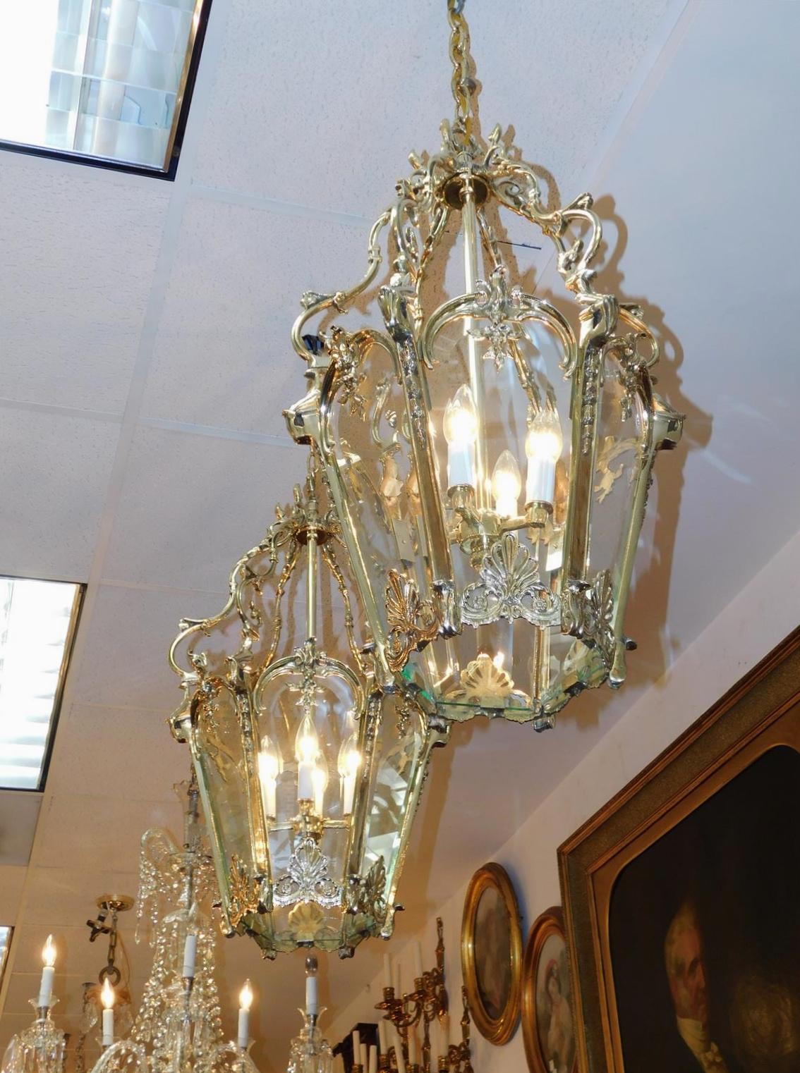 Pair of French brass hanging hall lanterns with decorative foliage scrolled frames, graduated bell flowers, shell accents, four light interior clusters, and retains the original six paneled beveled glass. Originally candle powered and have been