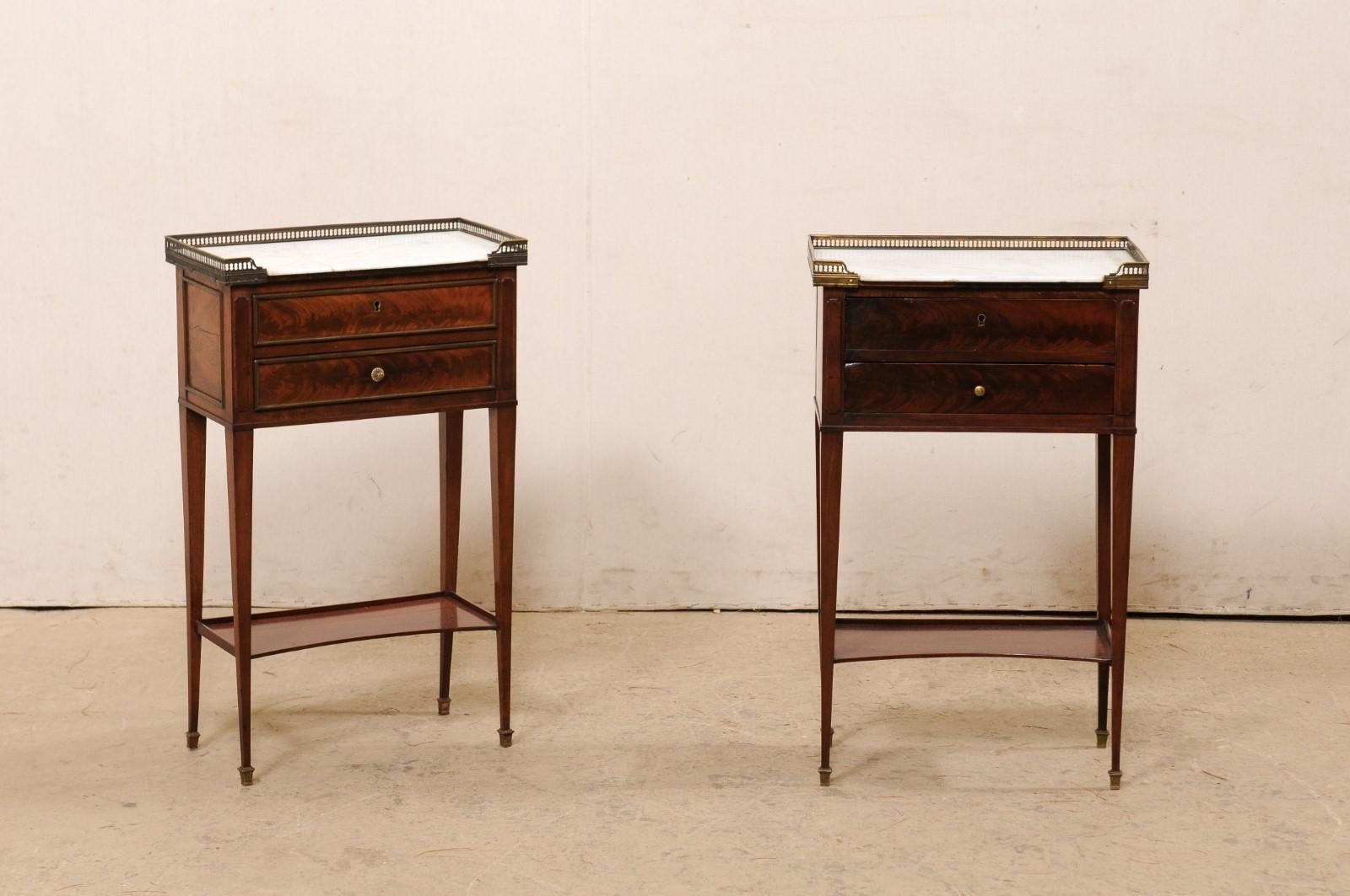A French near-pair of marble-top side chests with lower shelf, from the early 20th century. These antique commodes from France each feature a rectangular-shaped white marble top, surrounded with a raised brass gallery on three sides, atop a case