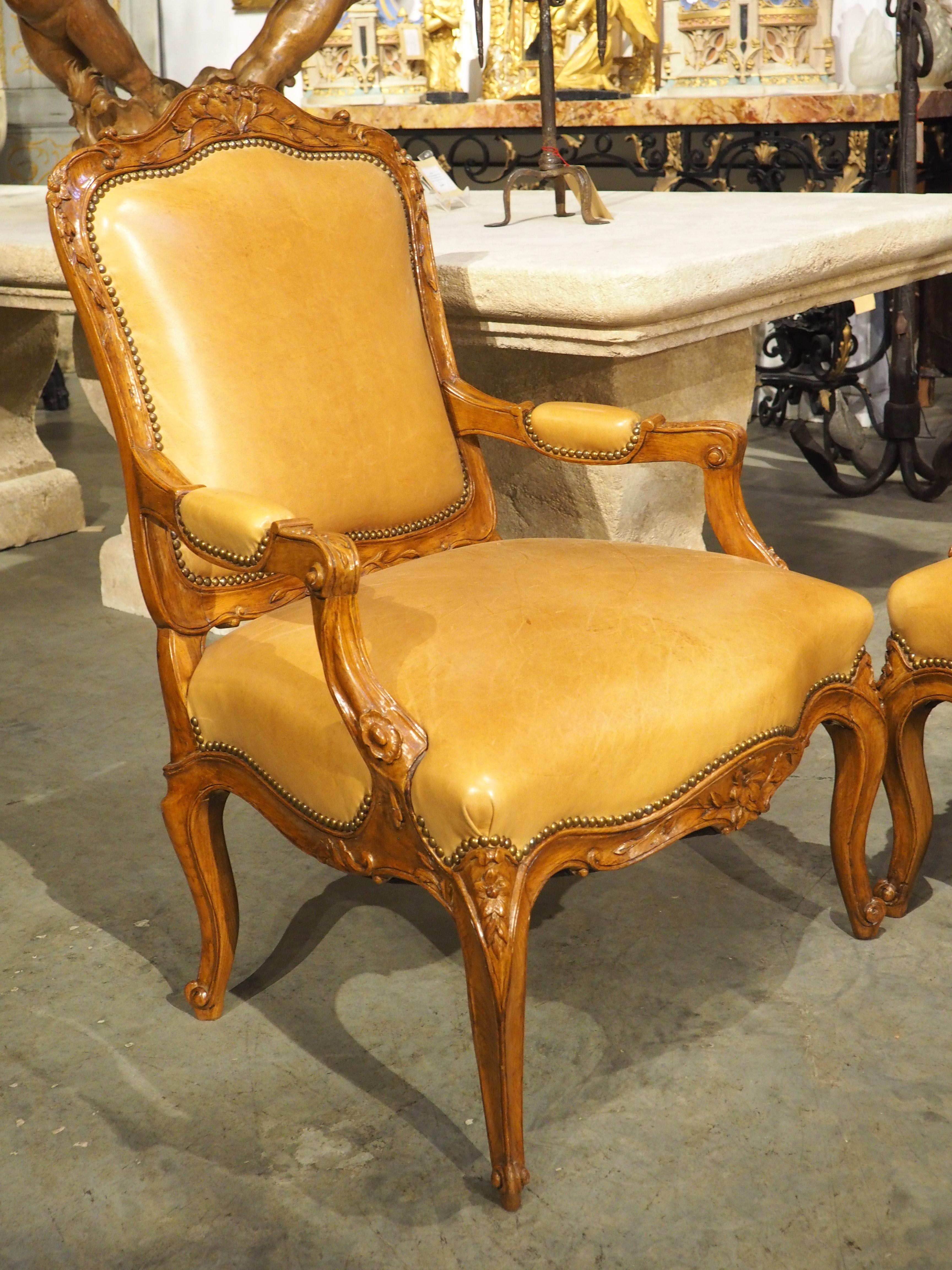 Hand-carved circa 1900 in the Regence style, this beautiful pair of armchairs have caramel-colored leather upholstery, forming a visually stunning palette with the rich brown patina of the varnished wood frames. Each chair back, manchette, and