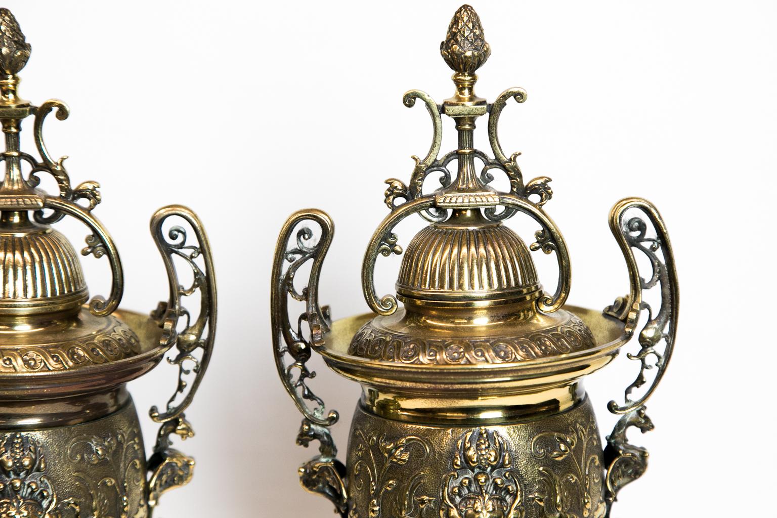 Pair of French Cassolettes has raised floral arabesques on a stipled background and are on a rouge de fer base. The removable top has a reeded dome topped with a pineapple finial.