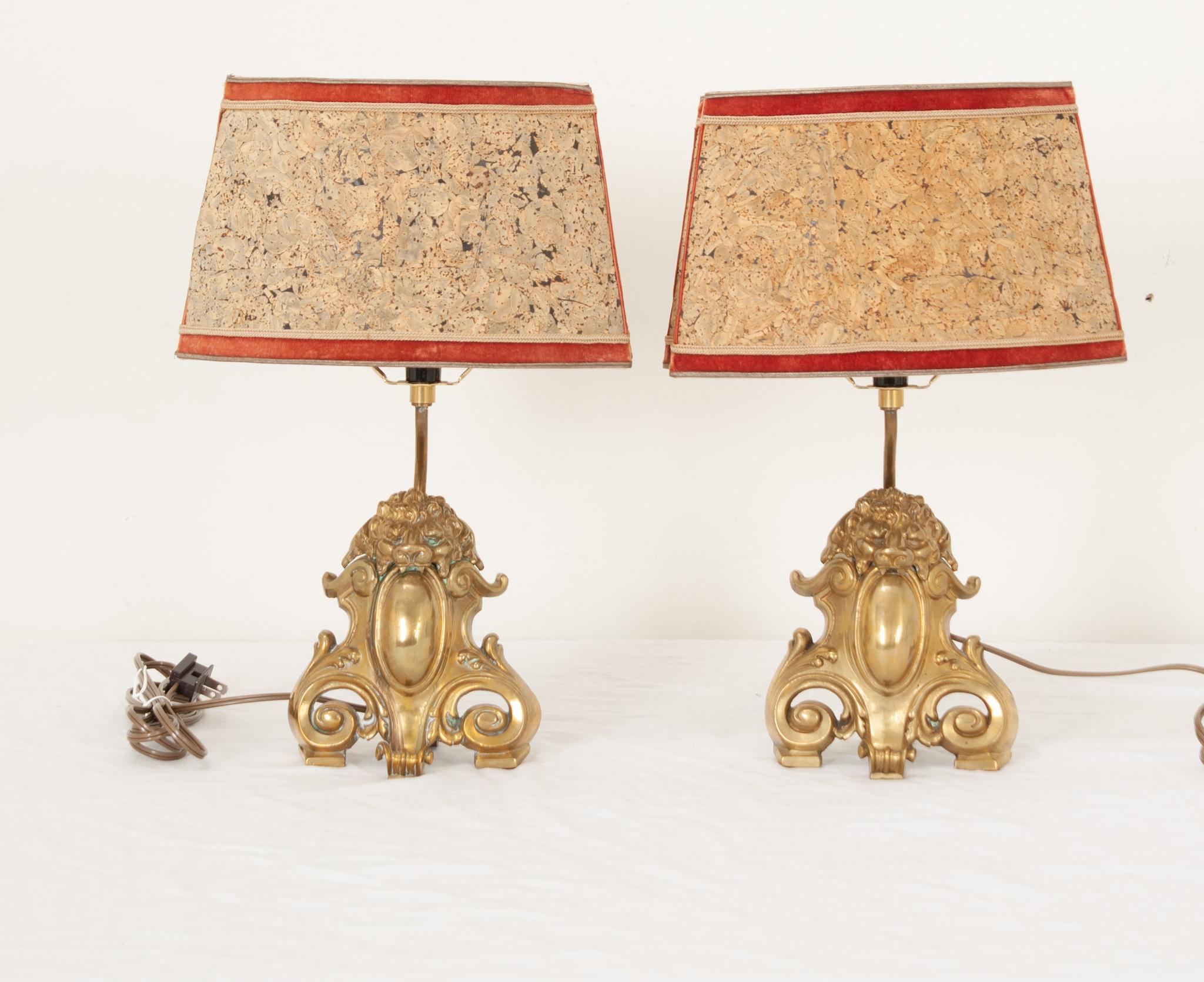 A superbly decorative pair of vintage French table lamps circa 1960. Eye-catching and dramatic in design, this petite set features brass bases of ferocious lions sinking their teeth into Rococo style ornaments featuring scrolling acanthus leaves.