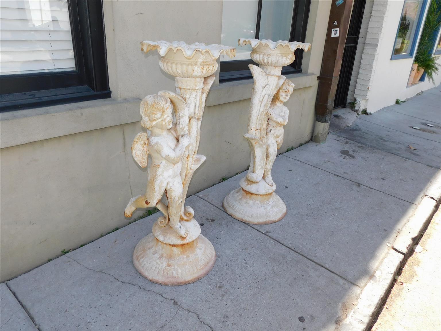 Pair of French cast iron painted garden torchieres with flanking standing cherubs, foliage gadrooned urns, central lily fluted columns with cat tails, and resting on circular gadrooned fluted bases, Early 19th century