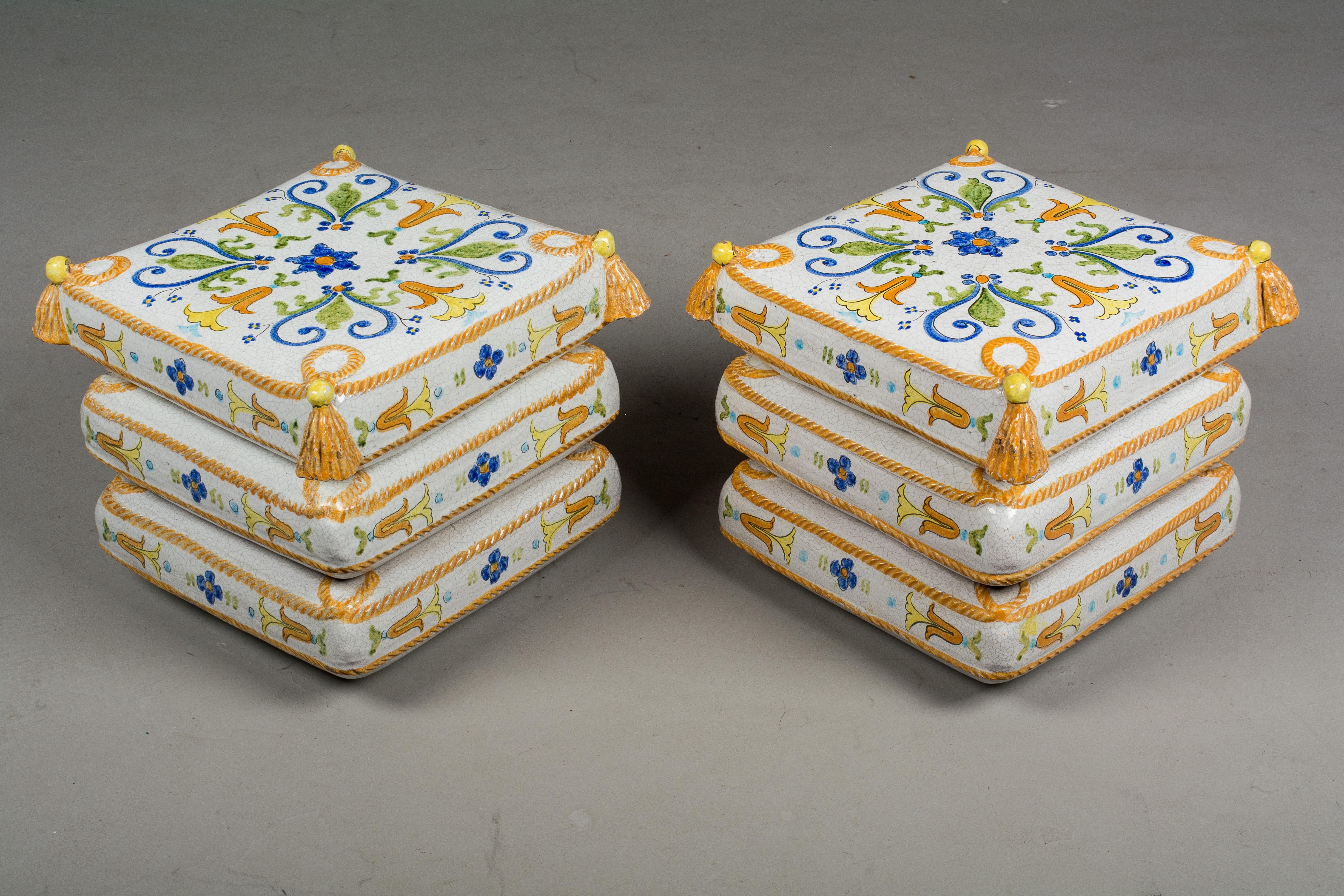 A pair of French ceramic garden stools, each made to look like a stack of three upholstered cushions with rope trim and tassels. White craquelure glazed ground with hand painted floral decoration in bright blue, yellow, green and gold. Each weights