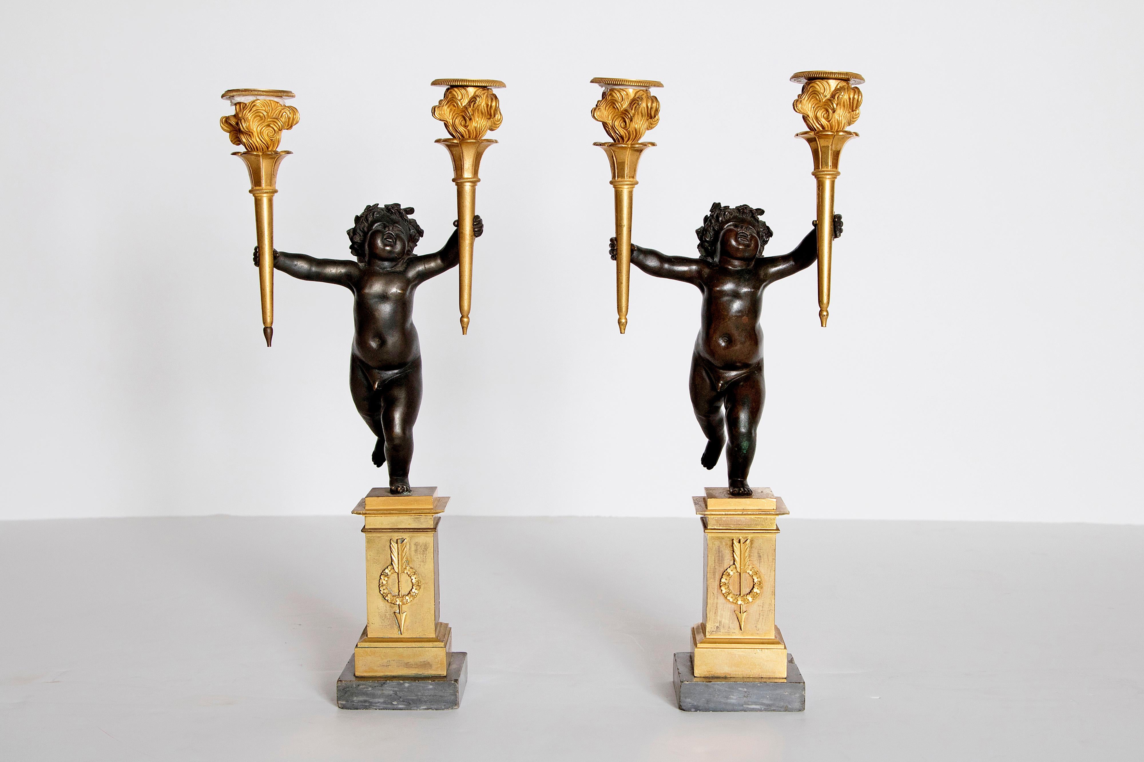 A fine pair of figurative candelabras in the form of a patinated bronze cherub running with a gilt torchiere in each hand. The cherubs stand on a gilded square pedestal base with an applied gilt wreath with an arrow. The cherubs separate from the
