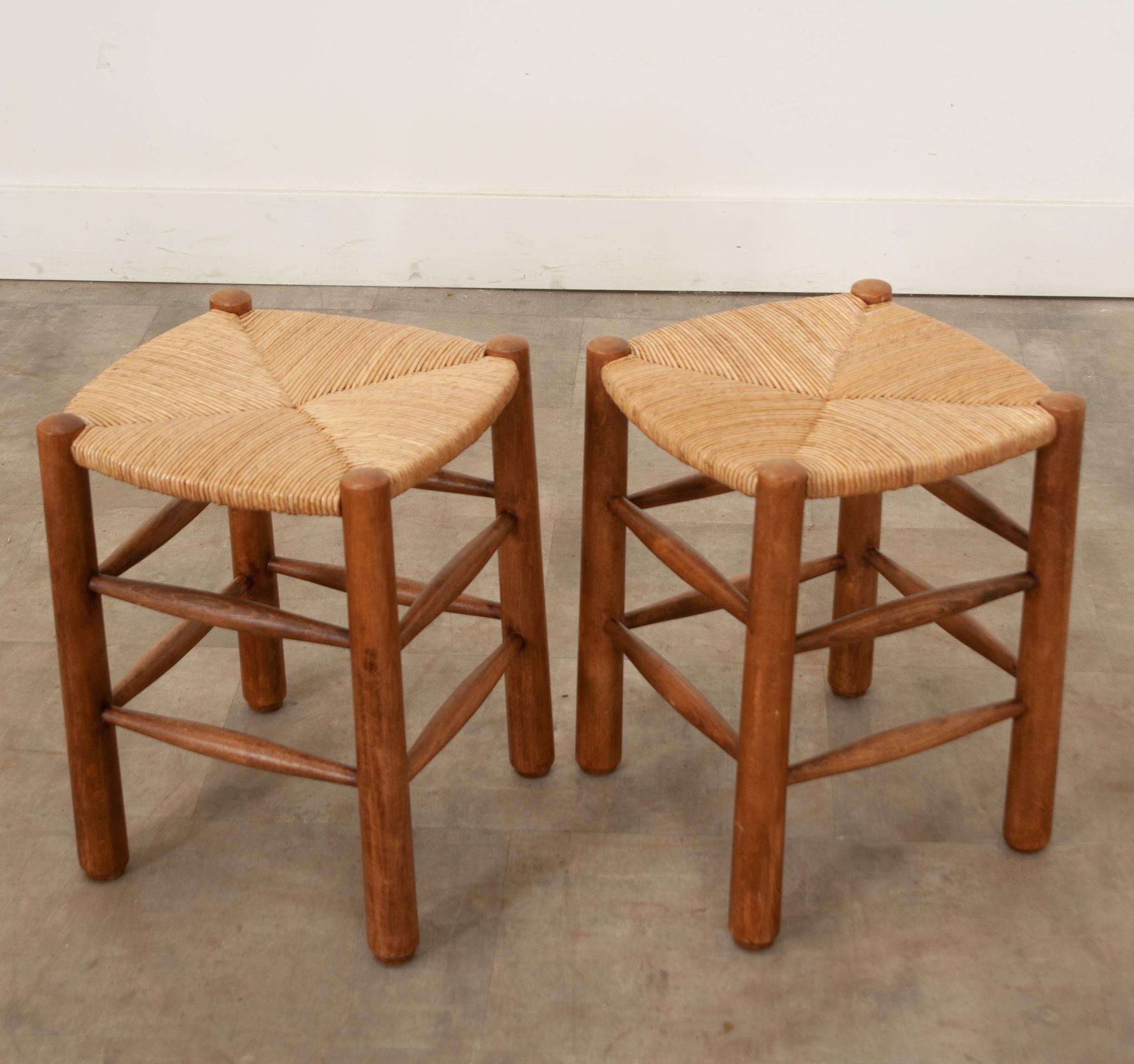 A vintage pair of rustic French rush seat & walnut stools in the style of Charlotte Perriand. Hand-crafted in France at mid-century, these well made stools are comfortable and of solid wood construction with a rush woven seat. Sturdy tapering legs