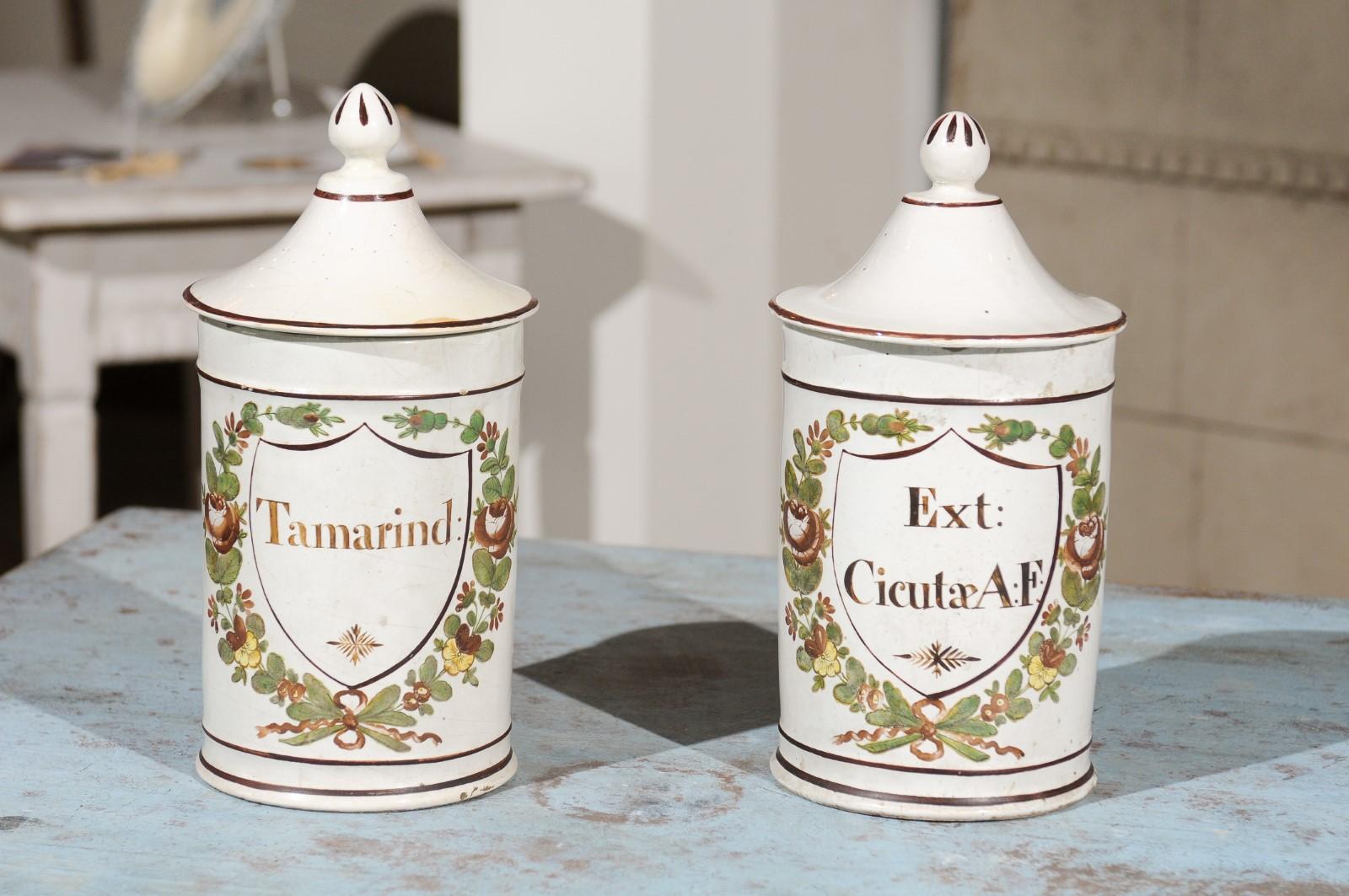A pair of French Directoire period ceramic lidded apothecary jars from the late 18th century, with hand-painted foliage motifs, labeled Tamarind and Ext. Cicutæ A:F, from the Deroche factory. Born in France during the Directoire, each of this pair