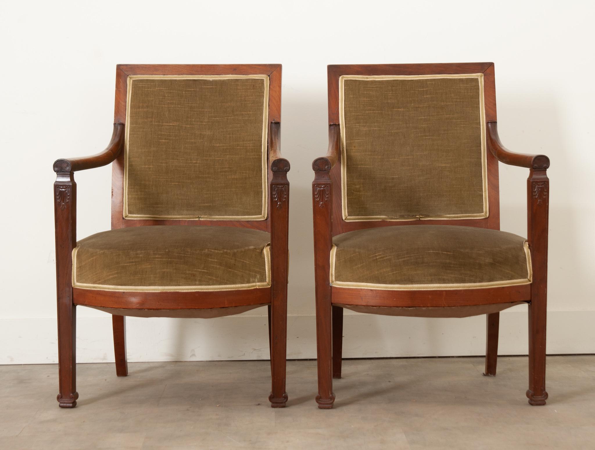 A handsome and sturdy pair of French Directoire square back fauteuils are tasteful and comfortable. These mahogany occasional chairs are upholstered in a worn green velvet and accented with gold tape trim. The arms are rounded and smooth and attach