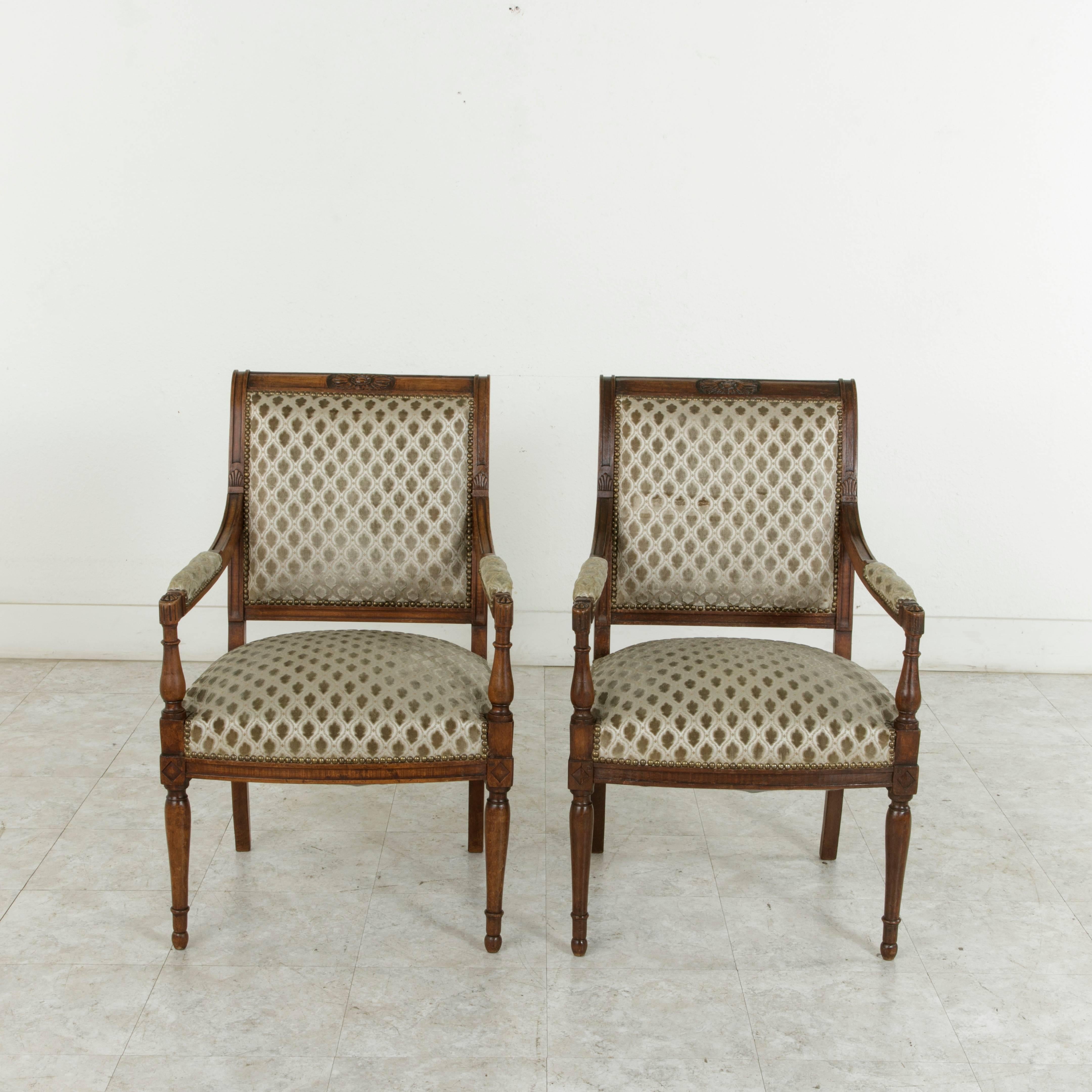 This elegant pair of French walnut armchairs from the Directoire period features detailed hand carvings of rosettes and stylized scalloped shells on the armrests, and an elongated central rosette at the crown. The turned legs of the piece meet the