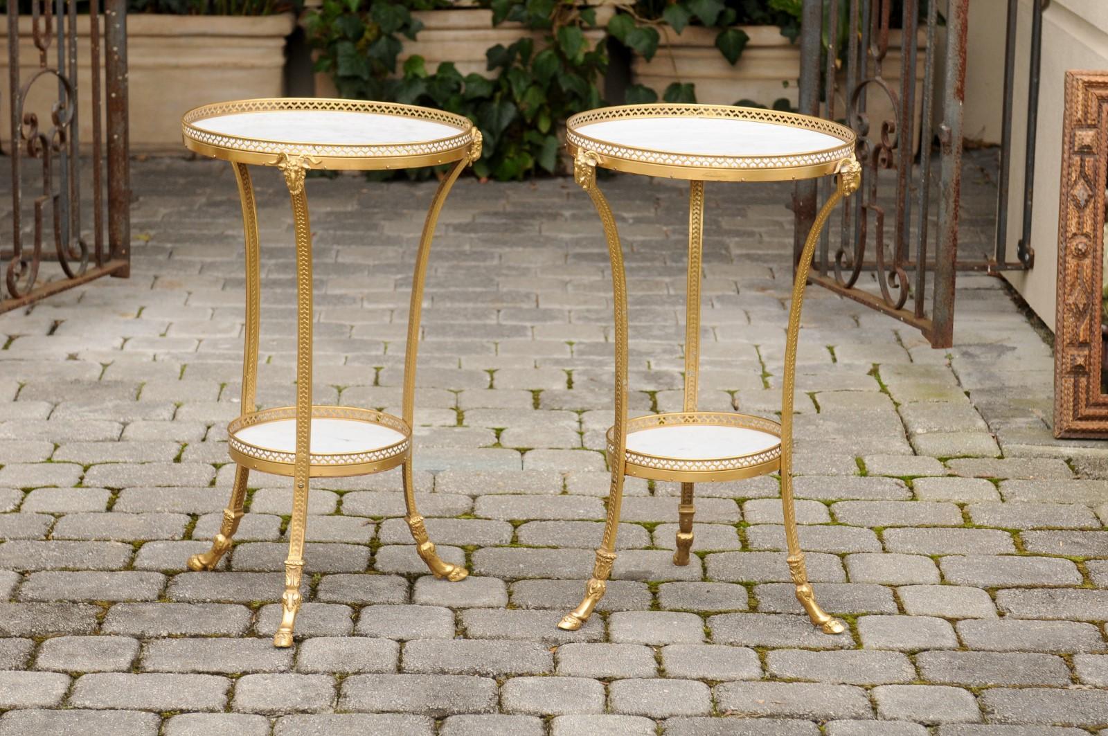 A pair of French Directoire style gilt bronze guéridon side tables from the early 20th century, with white marble tops and lower shelves, ram's heads and hoofed feet. Born in France during the Roaring Twenties, each of this pair of French guéridons