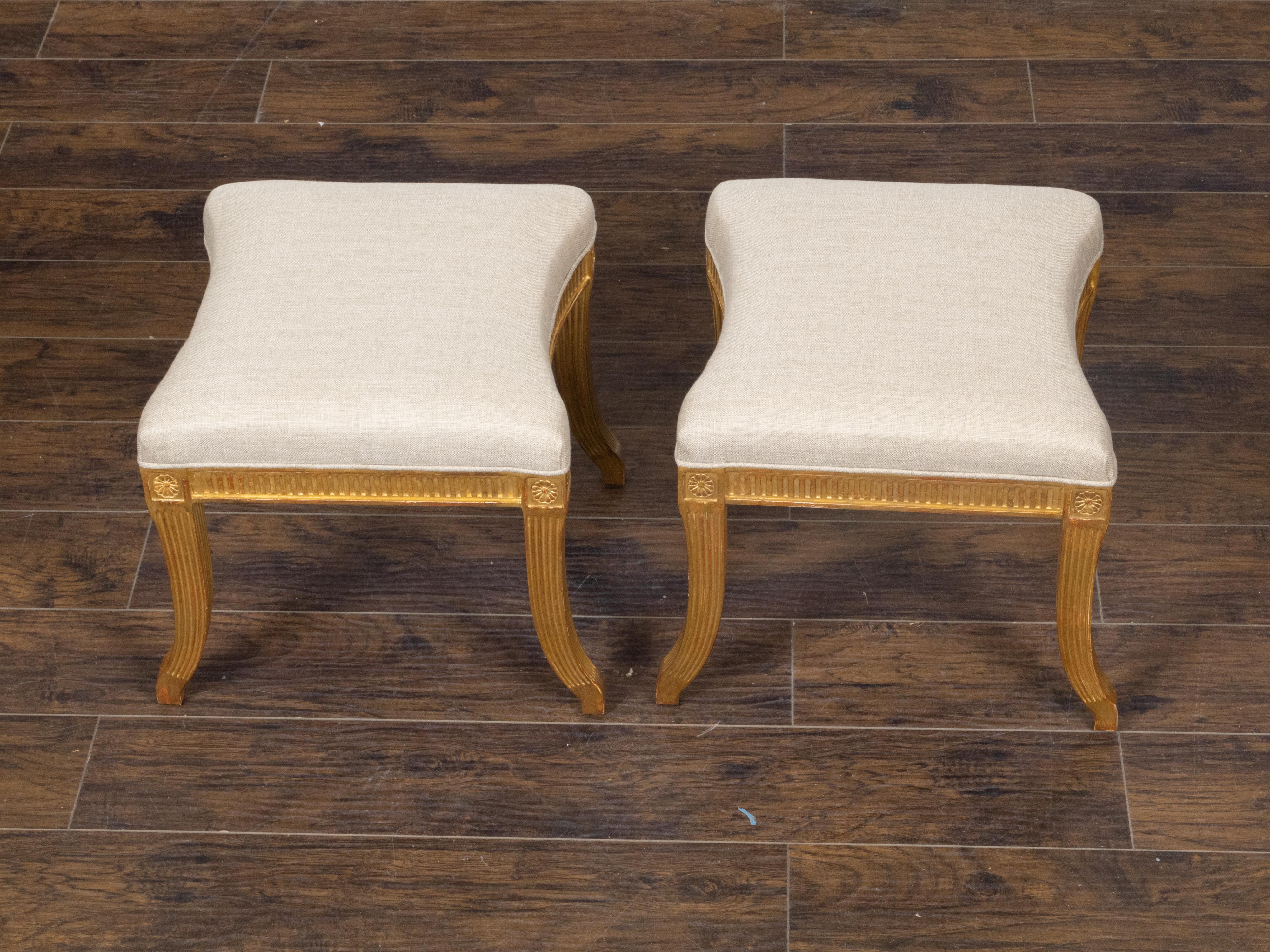 A pair of French vintage Directoire style giltwood stools from the mid 20th century with carved rosettes, fluted accents, saber legs and new linen upholstery. This handsome pair of French vintage Directoire style giltwood stools from the mid-20th