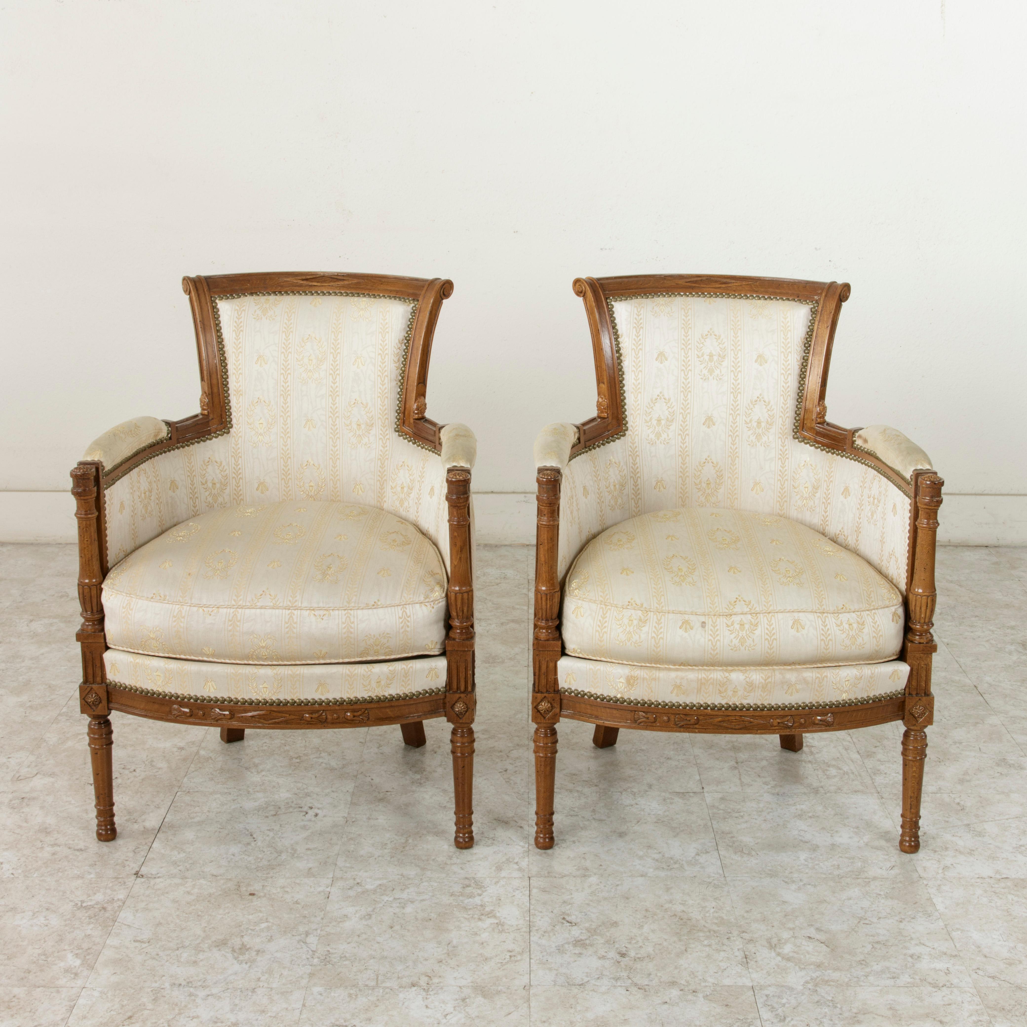 This pair of French Directoire style walnut bergères or armchairs from the turn of the 20th century features classic carved Directoire motifs of diamond shapes and rosettes. The armrests are detailed with fluted plinths and rosettes, and the front