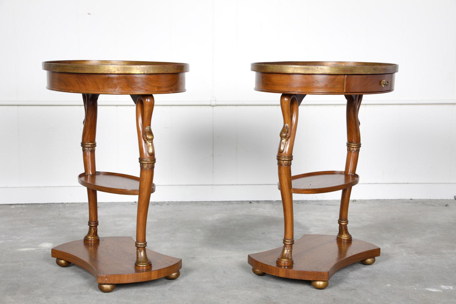 Elegant pair of 20th century walnut oval side tables with gilt accents in the style of French Directoire. The table’s frieze contains a paper-lined single drawer over figural swan carved legs. The table legs also support a shelf stretcher and