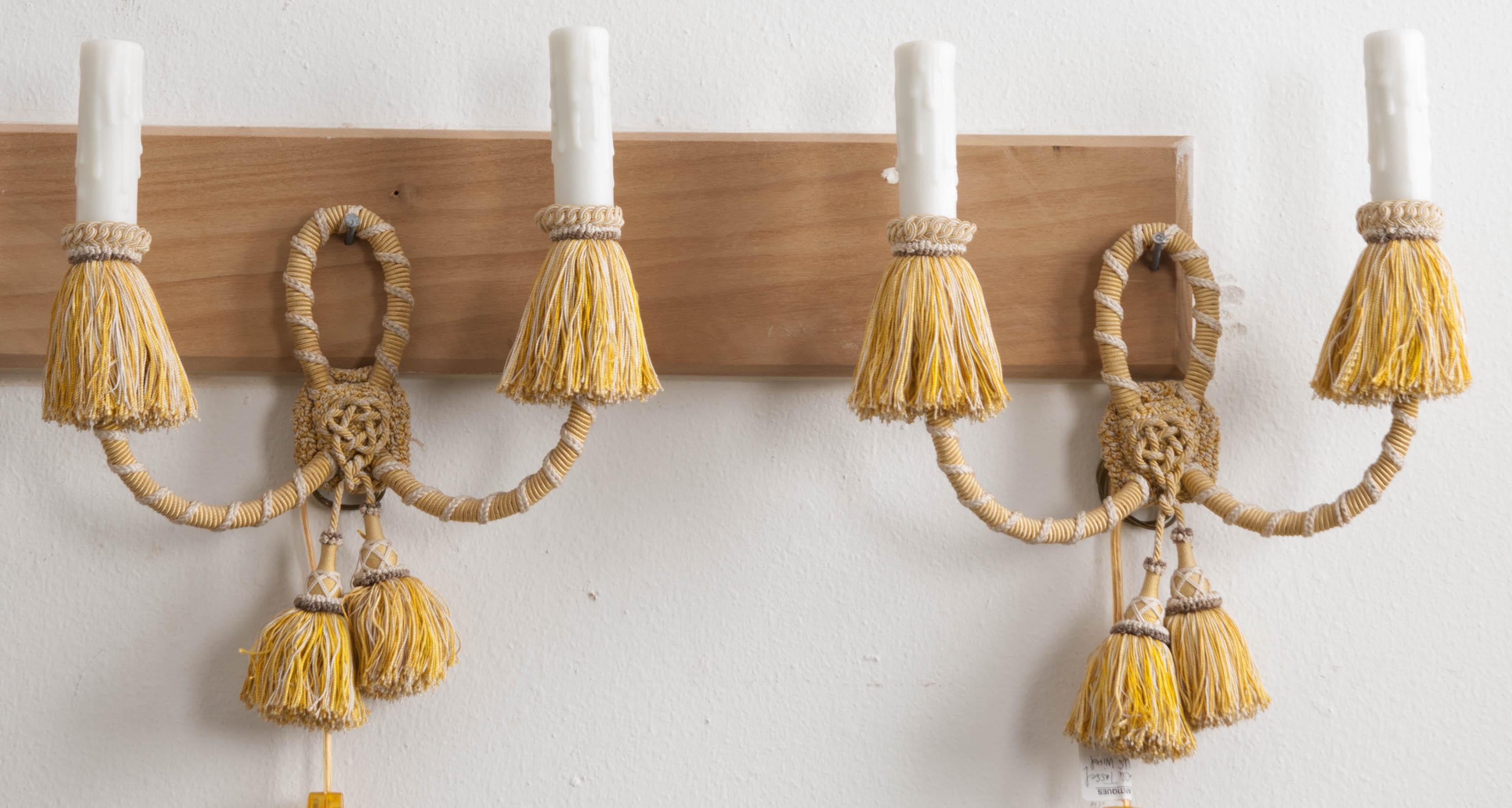 This fine pair of two-arm sconces, from France, circa 1900s, are covered in a pale gold and ivory cording with taupe accents. The faux candlesticks sit above a braided and fringed gimp and are mounted on upward scrolling arms joined by a central