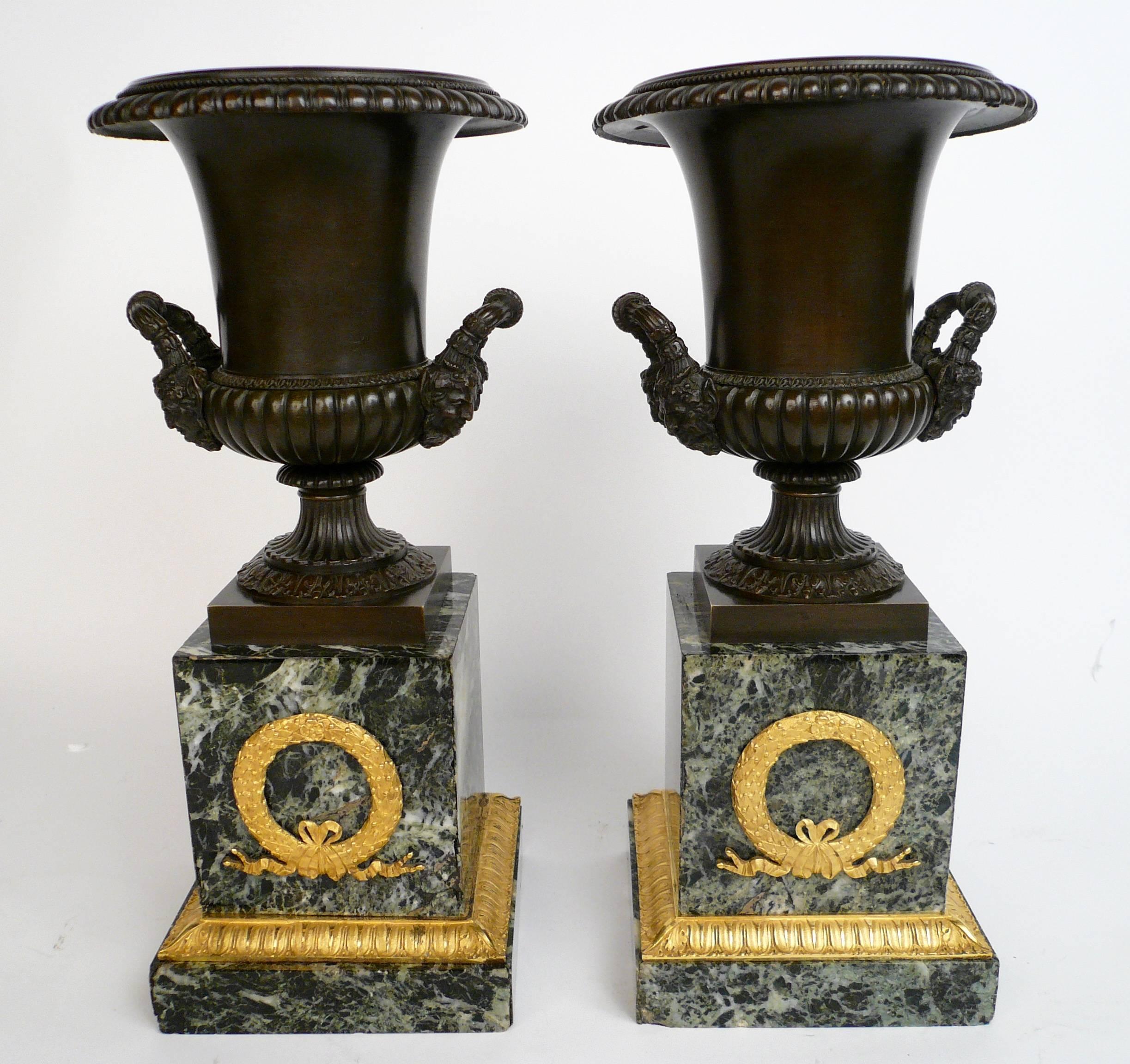 This handsome pair of urns feature neoclassical motifs, including acanthus leaves, egg and dart borders, and Bacchus mask handles. The Verde antico marble plinth bases are mounted with ormolu leaf and berry design wreaths.
 