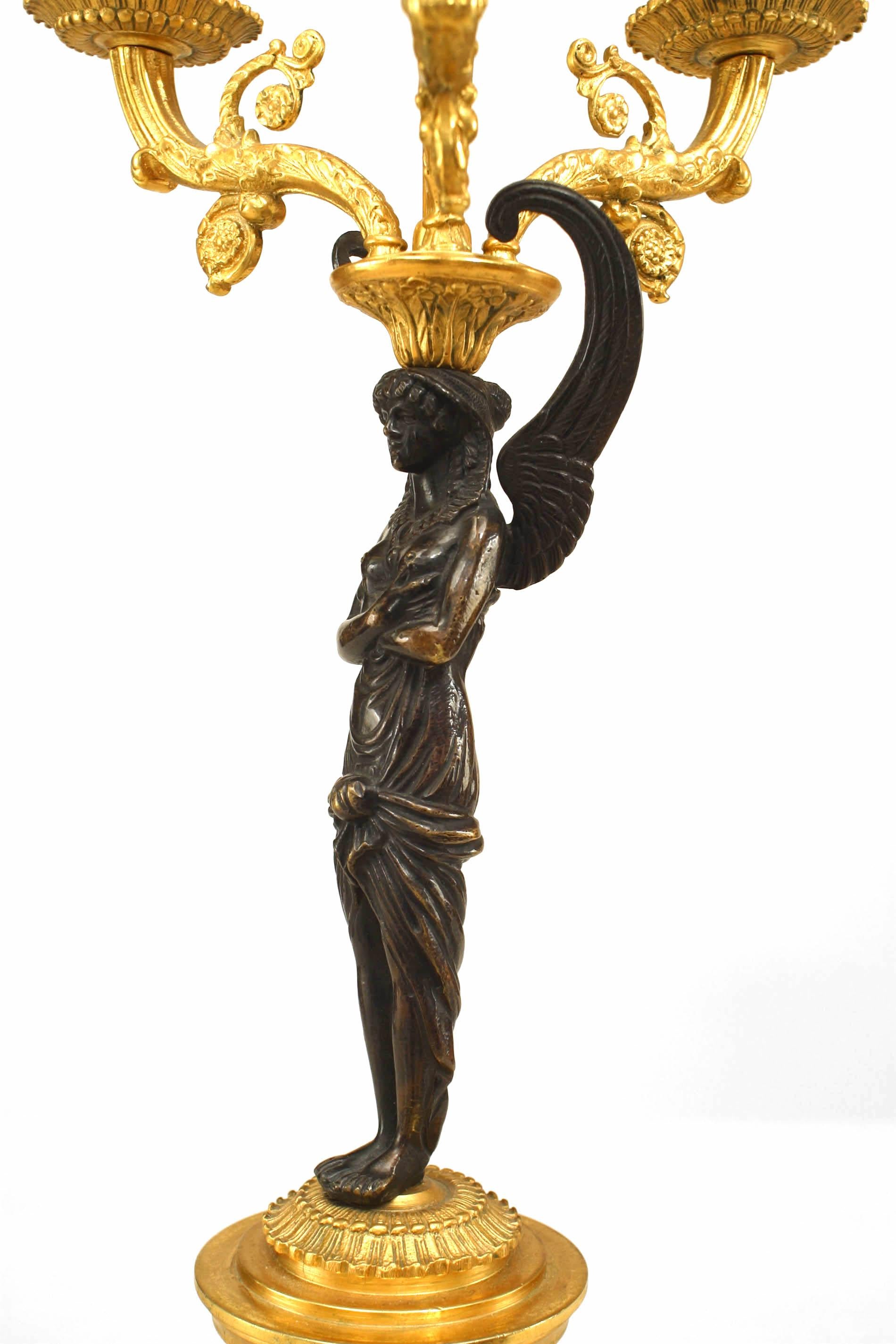 Paar vergoldete und patinierte 3-Licht-Kandelaber aus Bronze im französischen Empire-Stil (20. Jh.) mit geflügelten figuralen Säulen und Flammenabschluss auf ormolierter Basis. (Preisangabe als Paar).
 