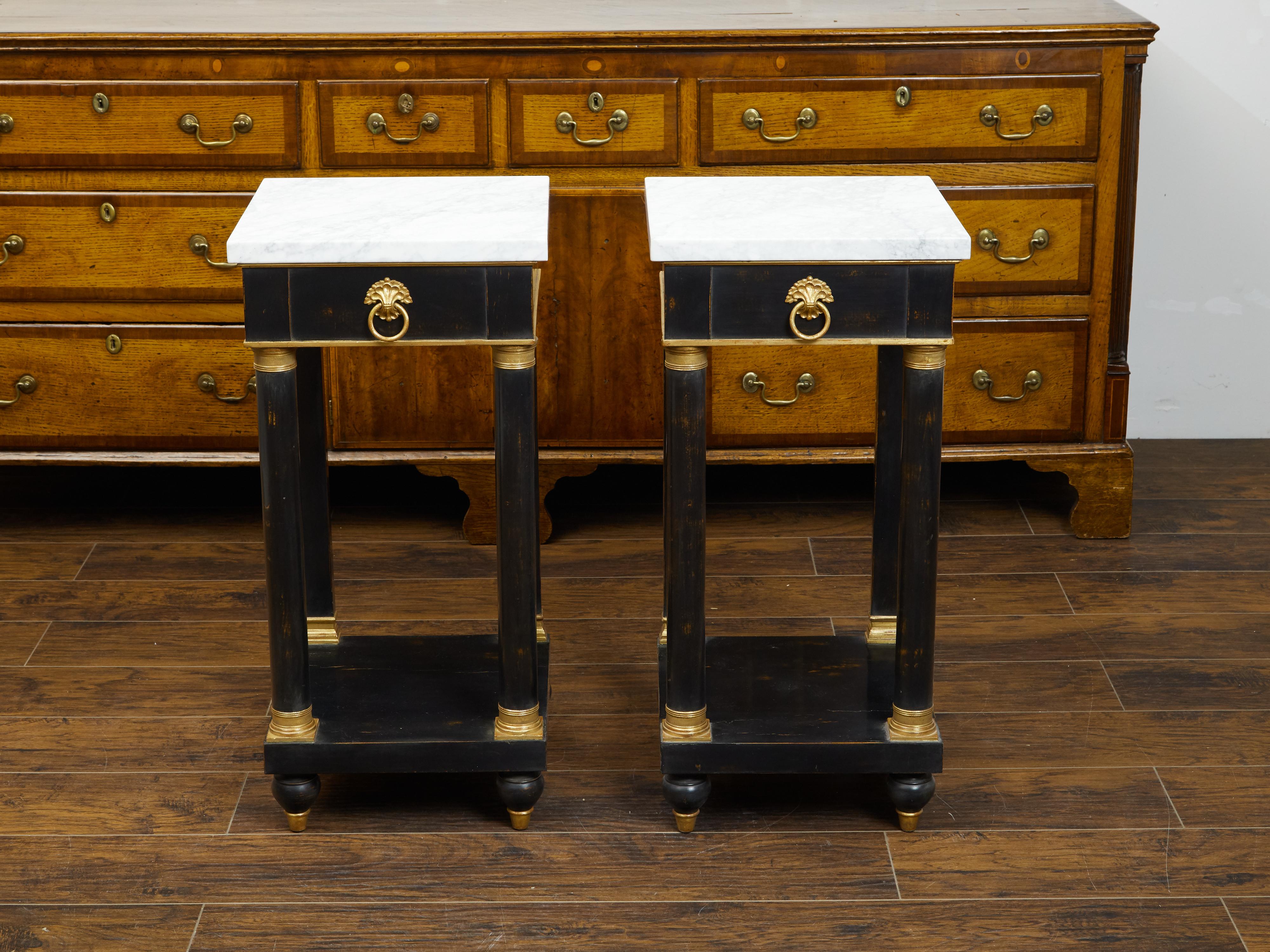A pair of French Empire style ebonized side tables from the mid 20th century, with marble tops and gilded accents. Created in France during the first half of the 20th century, each of this pair of tables features a square-shaped white veined marble