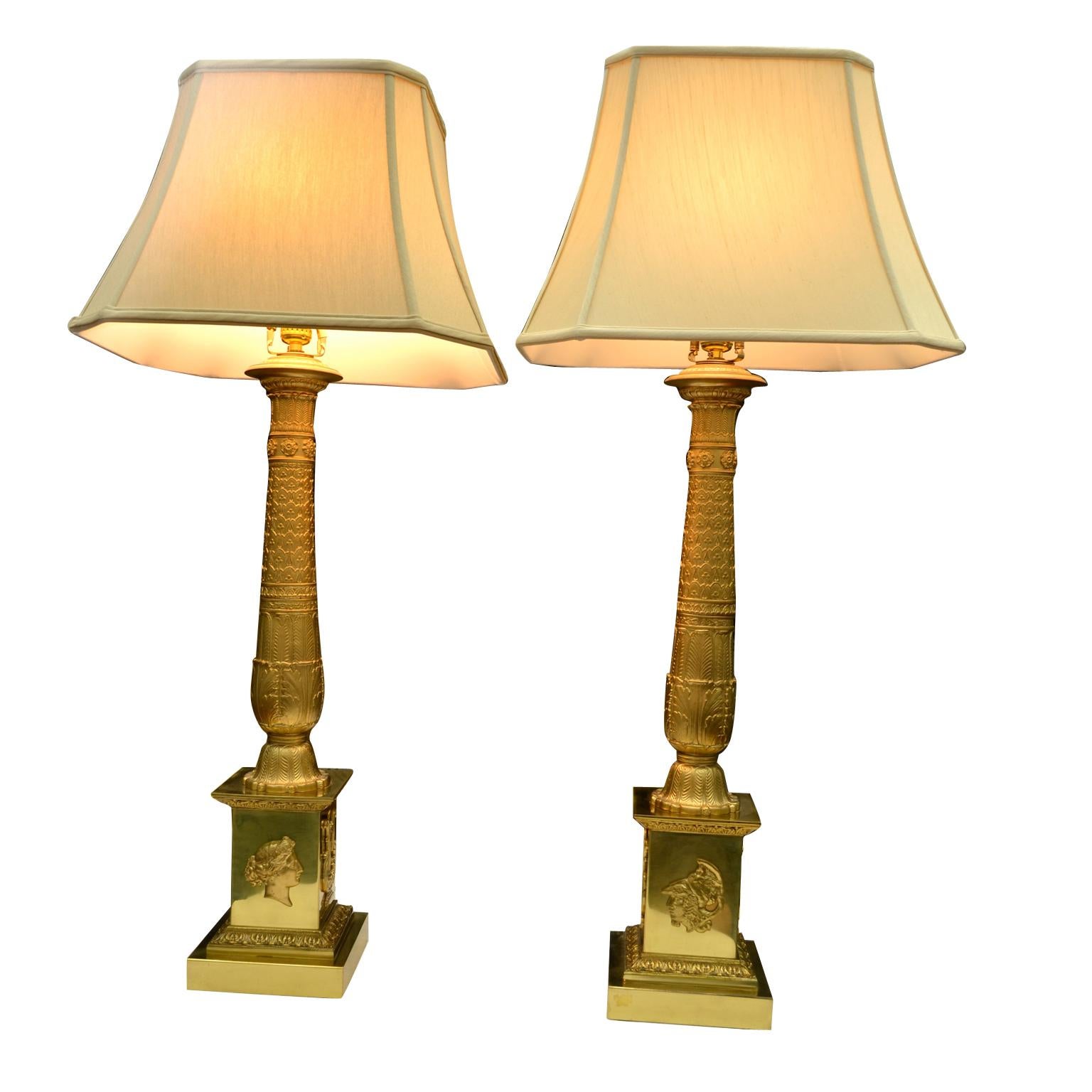 A pair of classical French Empire style gilded metal lamps (possibly earlier candelabra). The cast and decorated central round column sits on a stepped rectangular base, three sides of which are decorated with a lyre and a classical Roman profile,