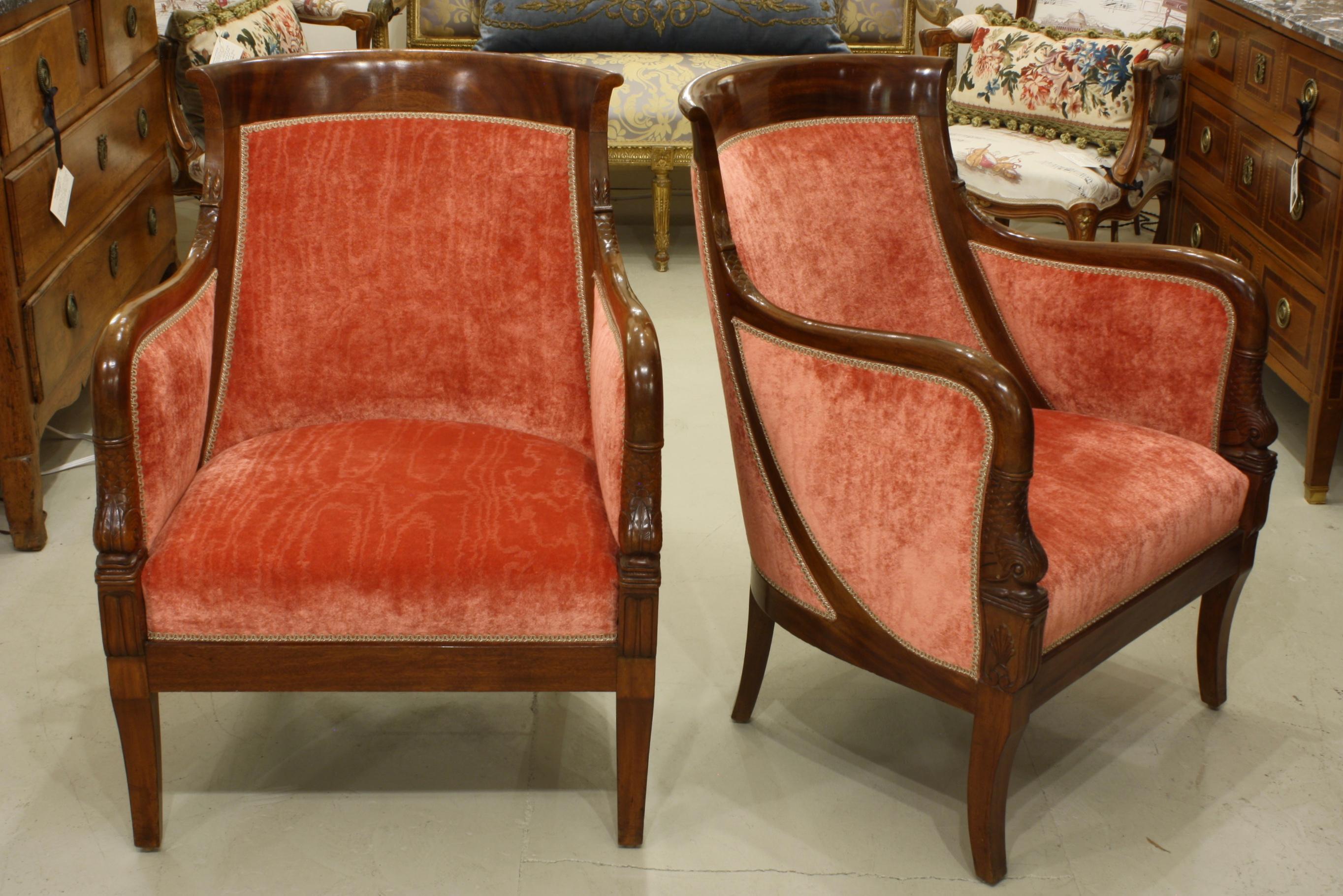 Pair of French Empire style mahogany bergeres or armchairs with carved mythical dolphin head on the arm ends and dolphin tails and scales where the arms meet the back (early 20th century). These chairs have been newly reupholstered in a gorgeous