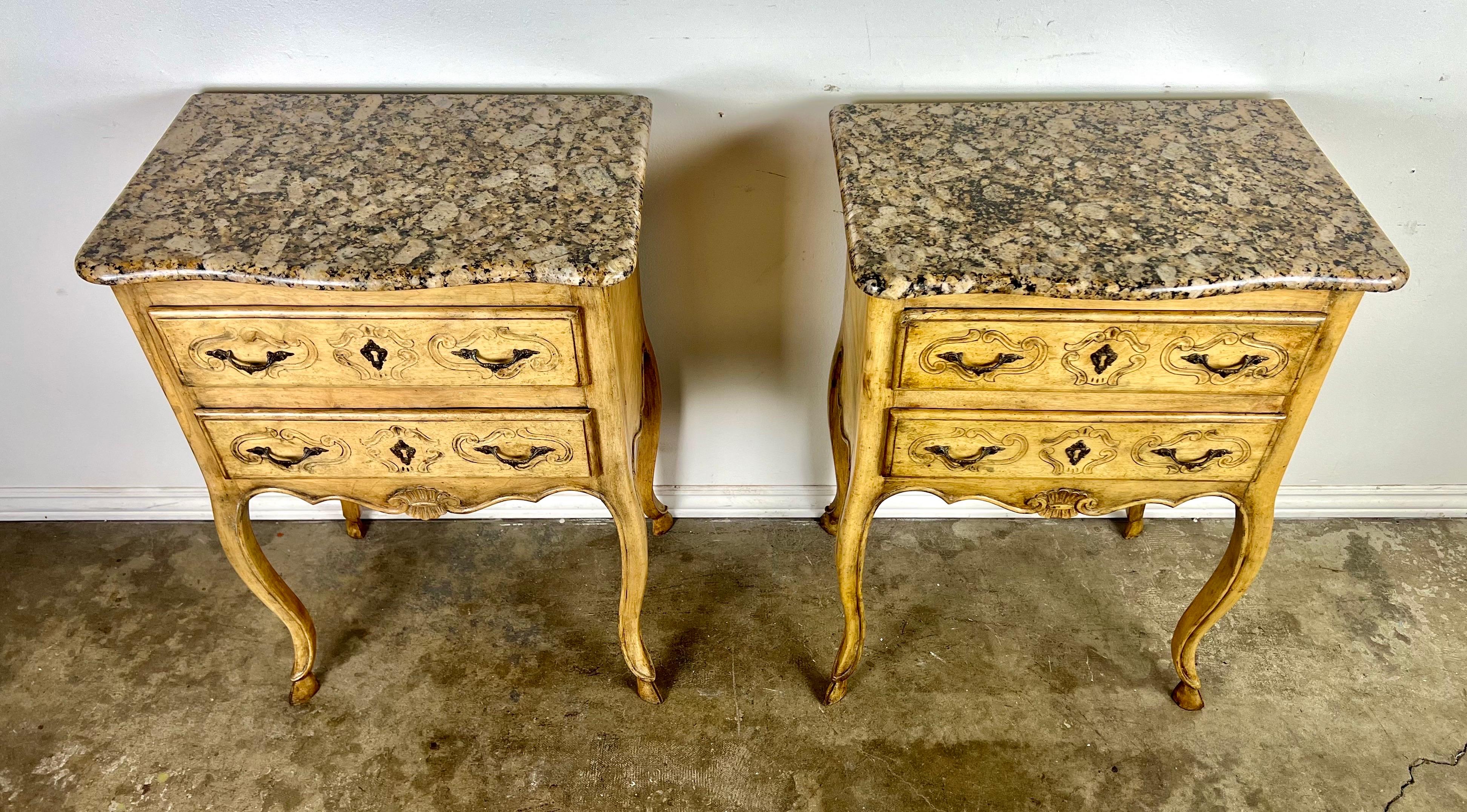 Pair of French Provincial painted side tables, each with two drawers and brass hardware, standing on cabriole legs ending in hoofed feet.  The granite tops feature a blend of cream, black, and touches of brown, beautifully complementing the tables'