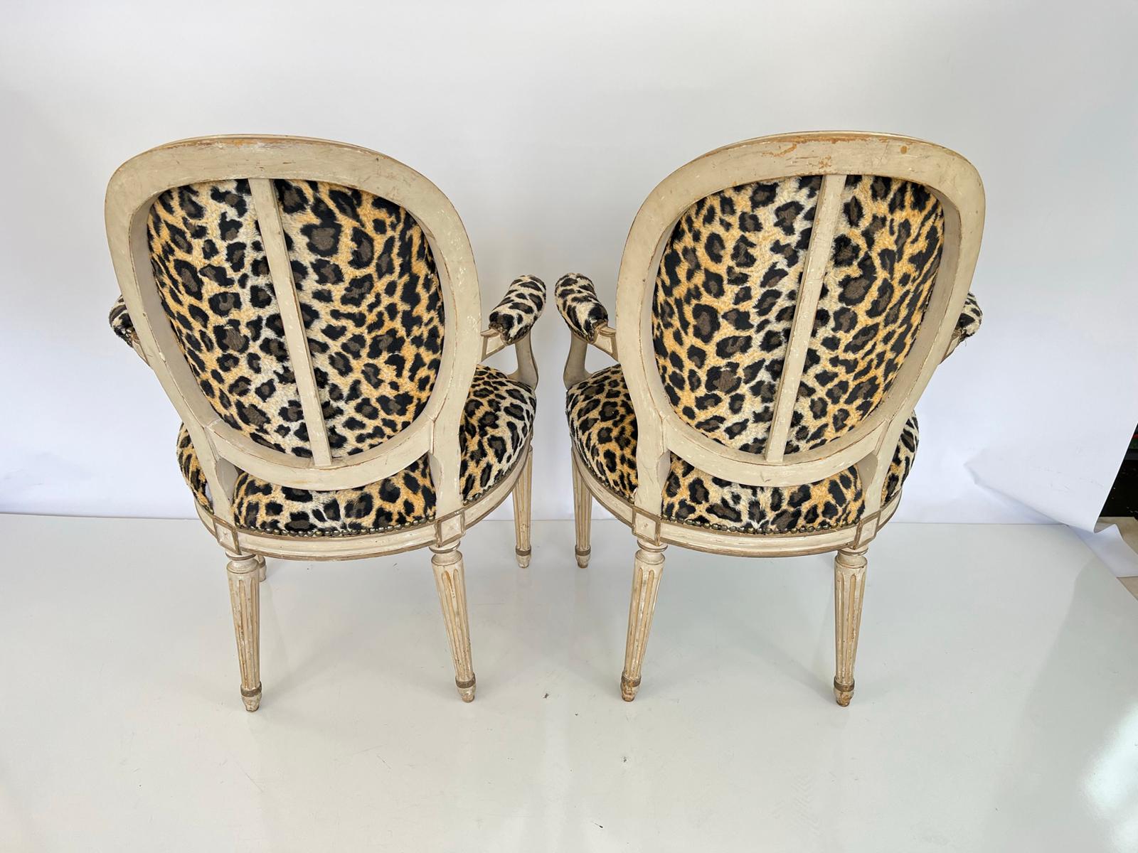 Pair of French armchairs, upholstered in faux leopard print with nailheads. Each chair having a painted, channeled frame, with oval, padded backrest. Its outswept arms with elbow cushions, supported by curvaceous terminals. A generous, crown seat is