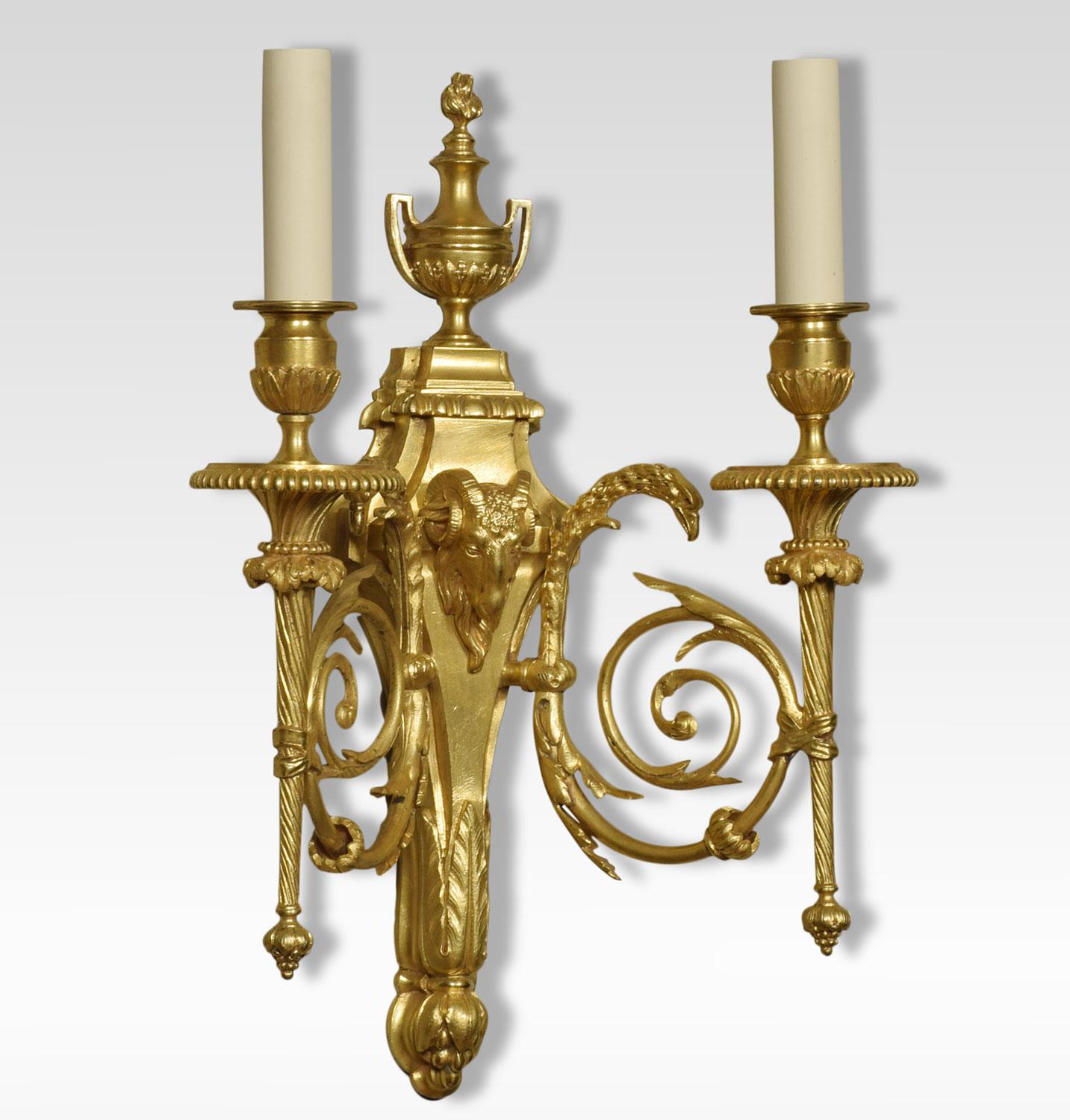 A pair of gilded bronze twin branch wall lights, the leaf scrolling arms with eagle-headed capitals having circular drip pans and acanthus sconces. Issuing from tapering backplate, with pierced finial above rams headed centre. The wall lights have