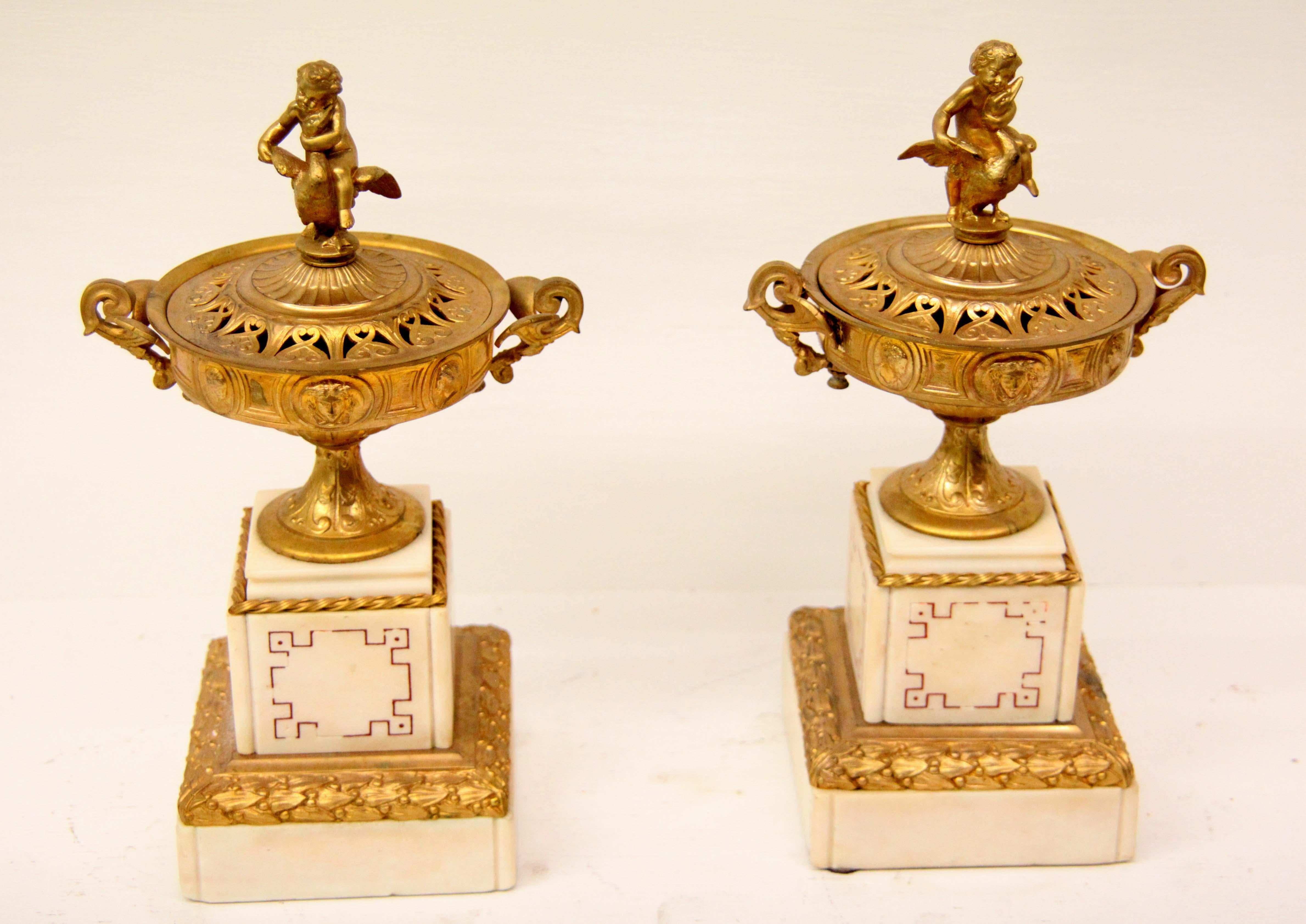 Pair of French gilt bronze and marble urns, the reticulated lid with a nymph and bird finial, the footed two handle urn with classical faces surrounding the perimeter, set on a four sided square marble plinth with geometric incised design, the