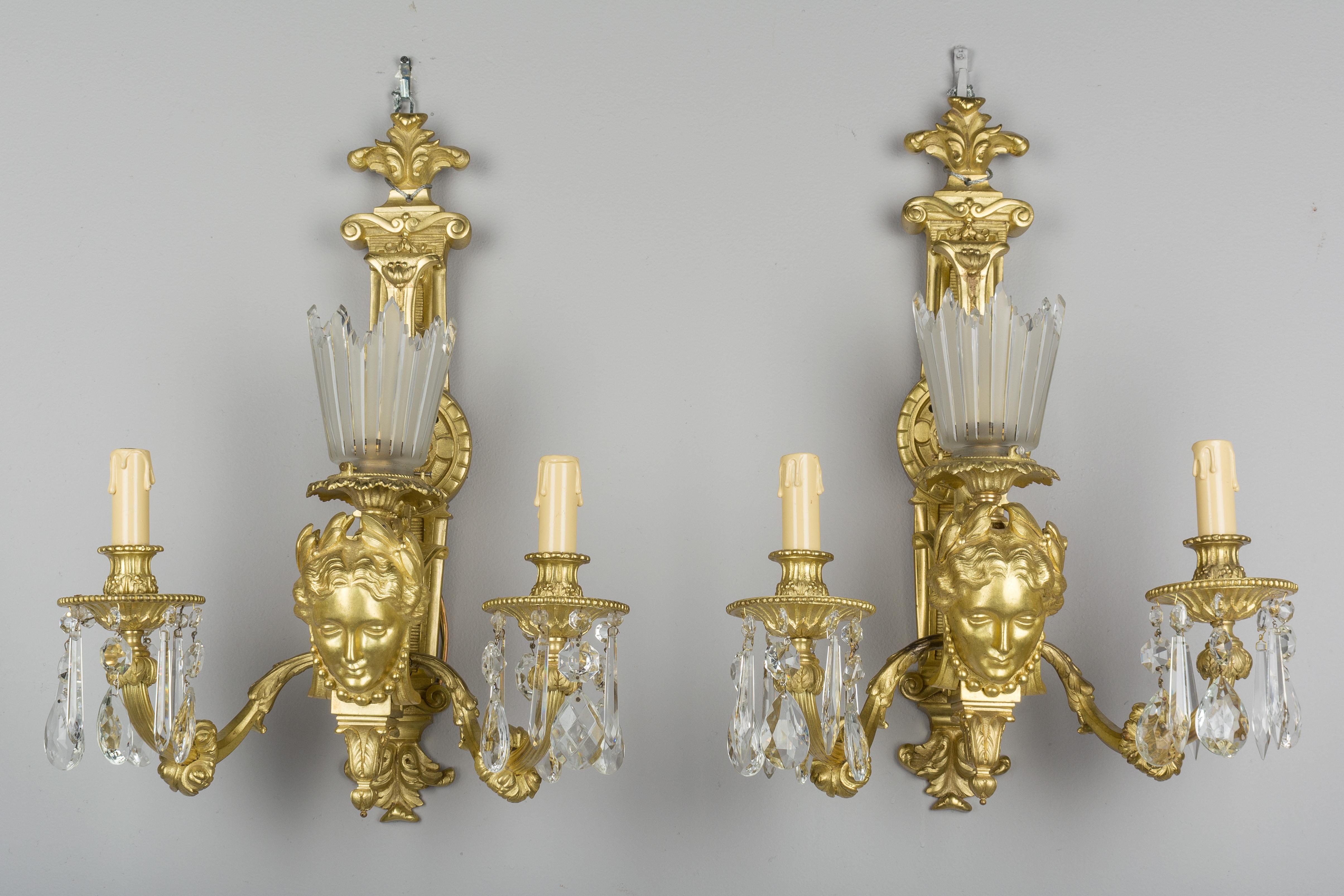 A pair of large heavy French gilt bronze three-light sconces, each with a classical woman's face below a crown-like frosted glass rayed shade and flanked by two candelabra with dripping crystal prisms. Fleur-de-lis bracket design. Fine quality