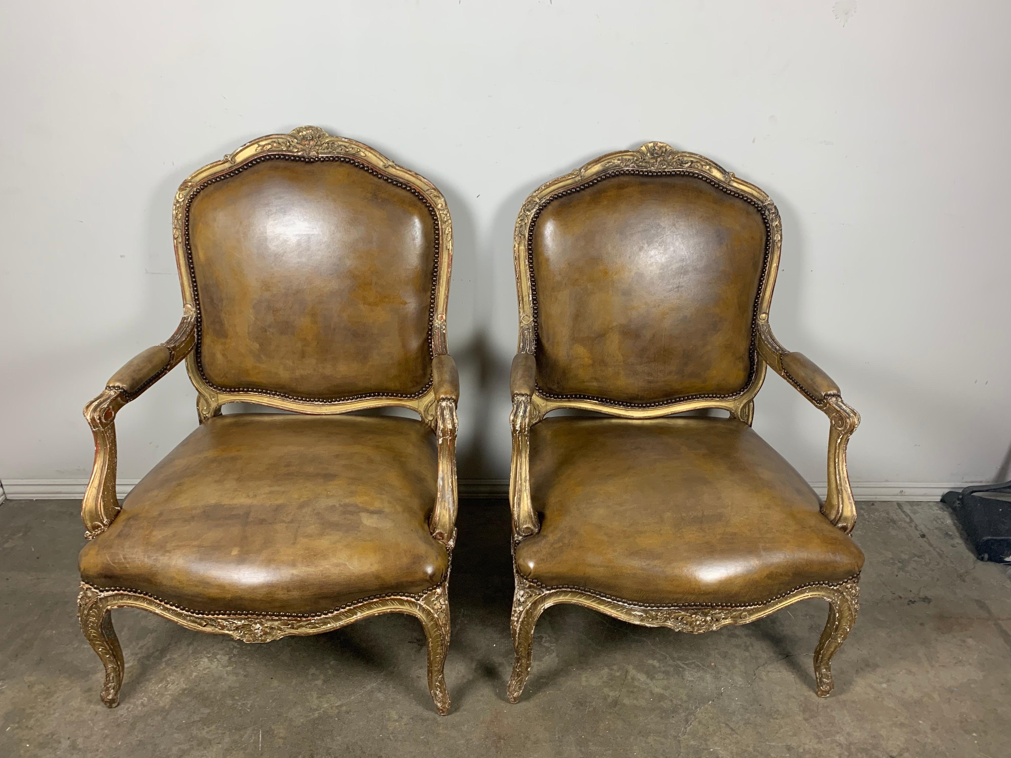 Pair of early 20th century French giltwood armchairs that are upholstered in a tobacco colored leather with nailhead trim detail. The chairs stand on four carved cabriole legs. Small carved flowers throughout. Distressed gilt wood finish.
