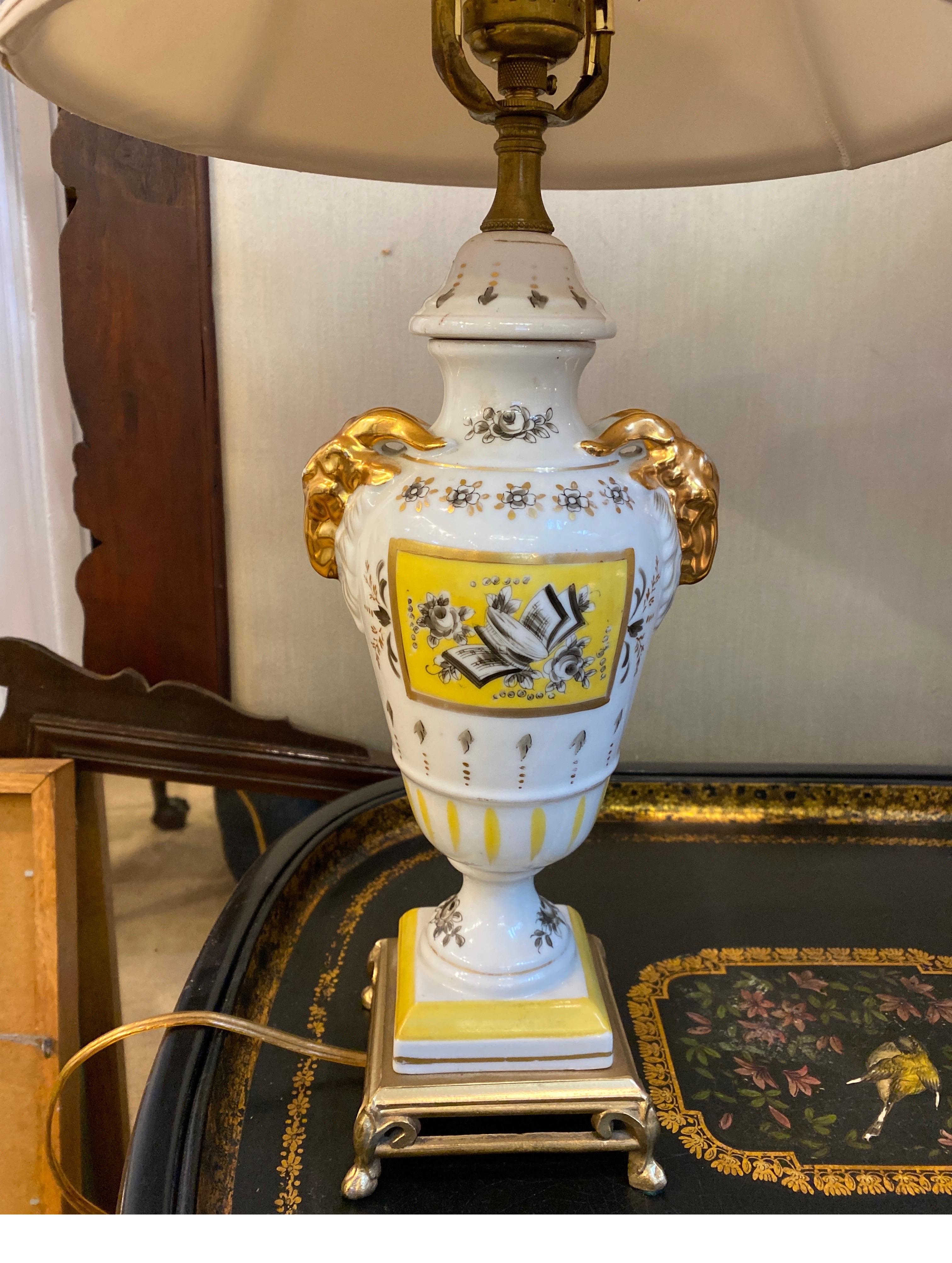 A pair of hand painted French porcelain lamps with gilt bases. The urn form with gilt rams head handles. These are a diminutive size, 20.5 inches tall to the top of a shade. These shades are for photographic purposes only and not included. 12.5