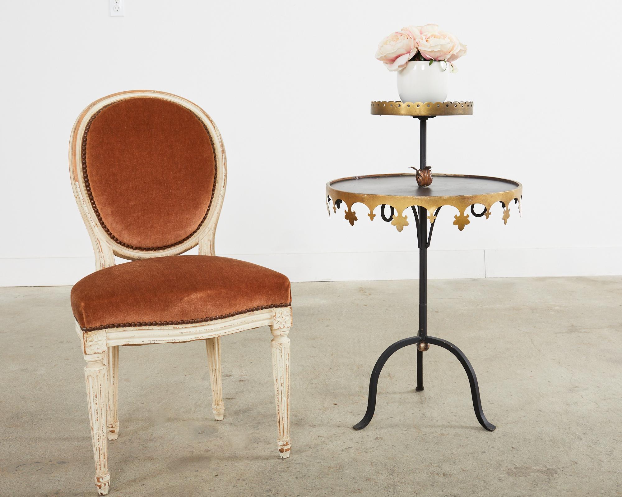 Whimsical pair of French style Hollywood Regency two-tier stands or drink tables featuring an iron tripod frame. Each stand has two tiers the bottom having a round 20 inch wide disk of tole with an inverted fleur de lys gilt border. The top tier is