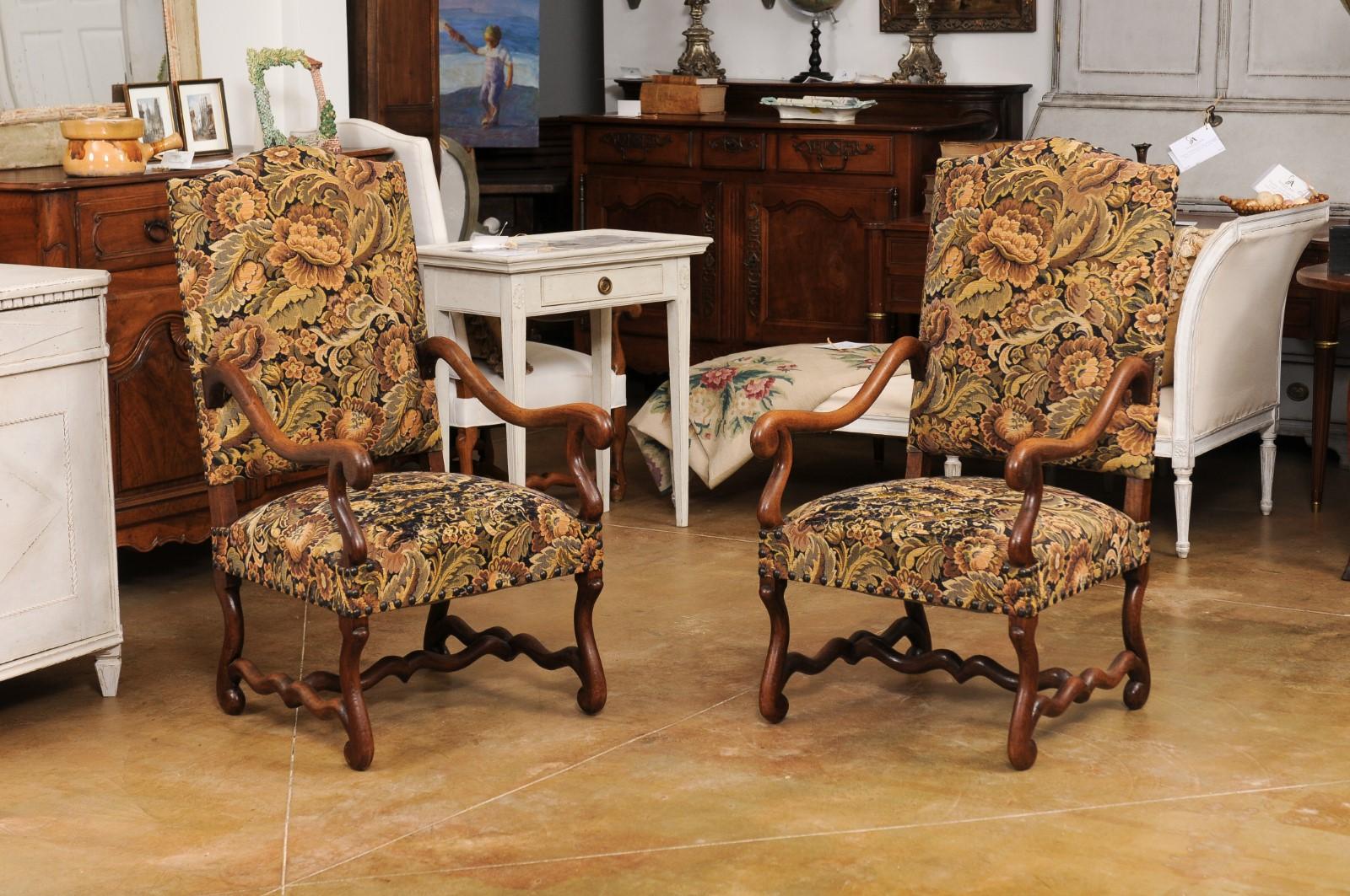 A pair of French Louis XIII style wooden os de mouton armchairs from the 19th century, with slanted camelbacks, large scrolling arms, cross stretchers and used upholstery. Created in France during the 19th century, this pair of fauteuils showcases
