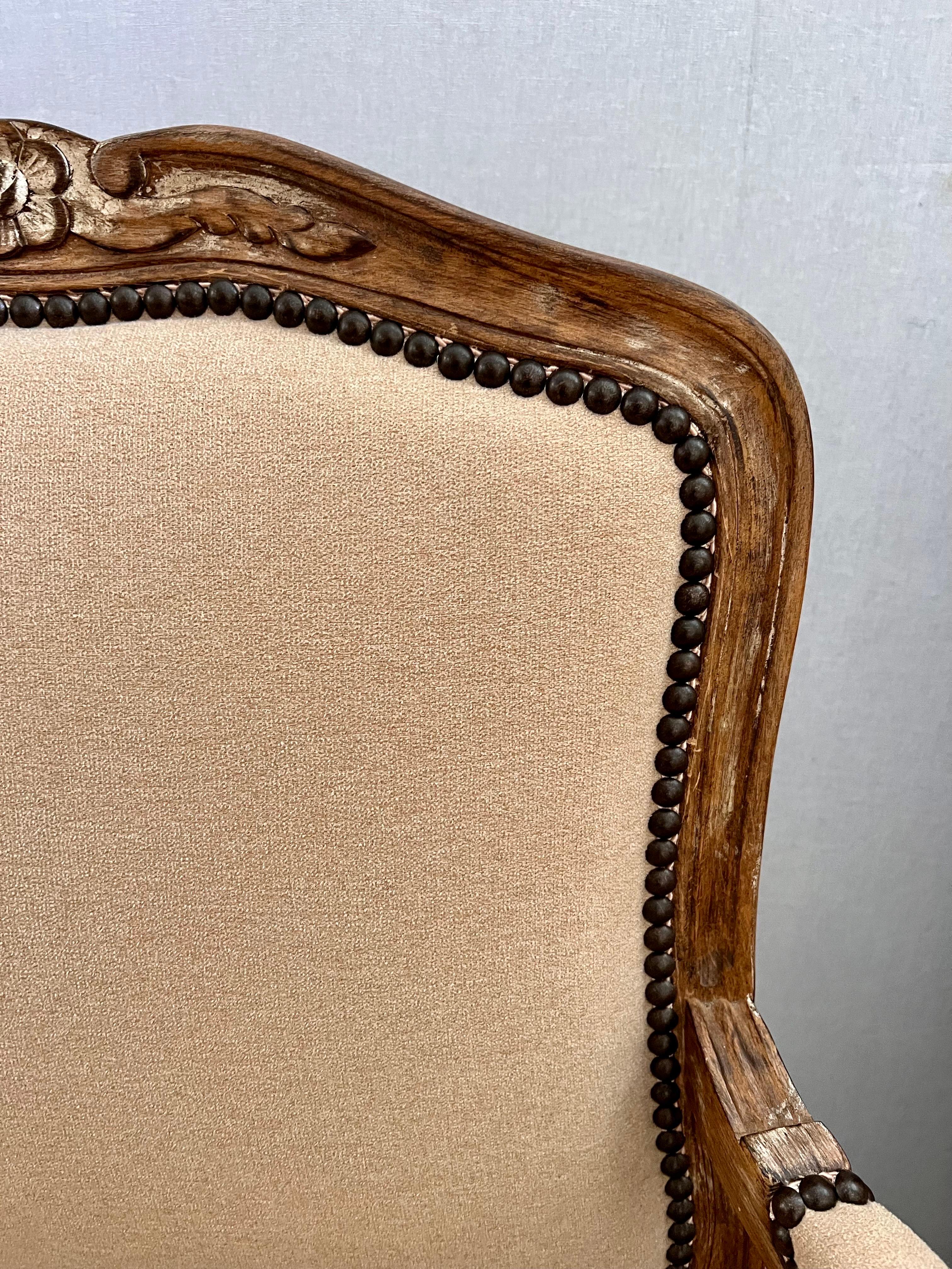 A superb pair of newly upholstered matching bergere chairs. They have natural wood carved frames with an intentionally distressed finish and are upholstered in a neutral beige soft thick fabric and finished with brass nailheads.