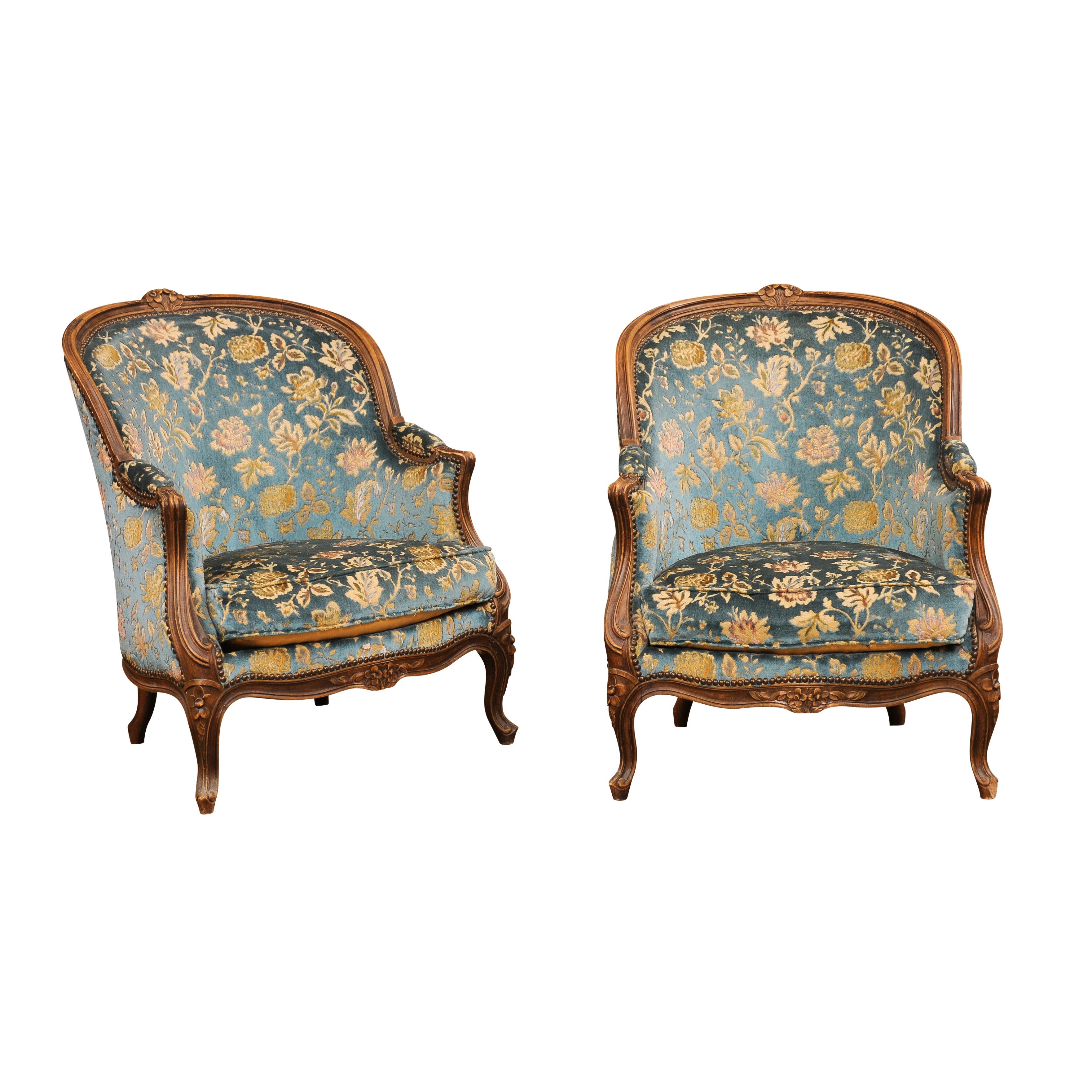 A pair of French Louis XV style walnut bergères chairs from the 19th century with carved crests, scrolling arms, floral carved apron, cabriole legs and floral upholstery. Enhance your living space with this exquisite pair of French Louis XV style