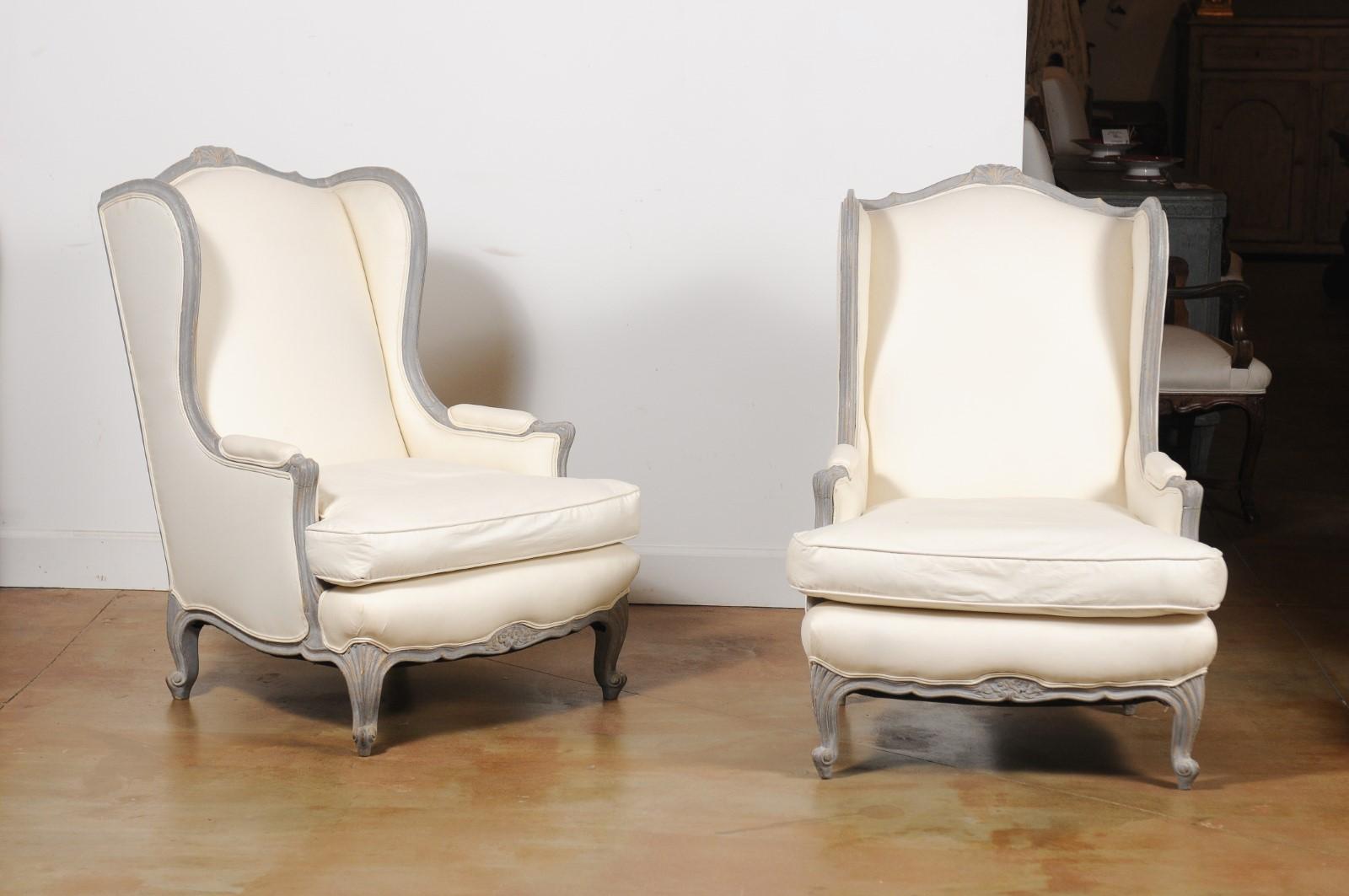 A pair of French Louis XV style painted bergères à oreilles from the late 19th century, with grey painted finish and upholstery. Created in France during the last quarter of the 19th century, each of this pair of bergères features a slanted