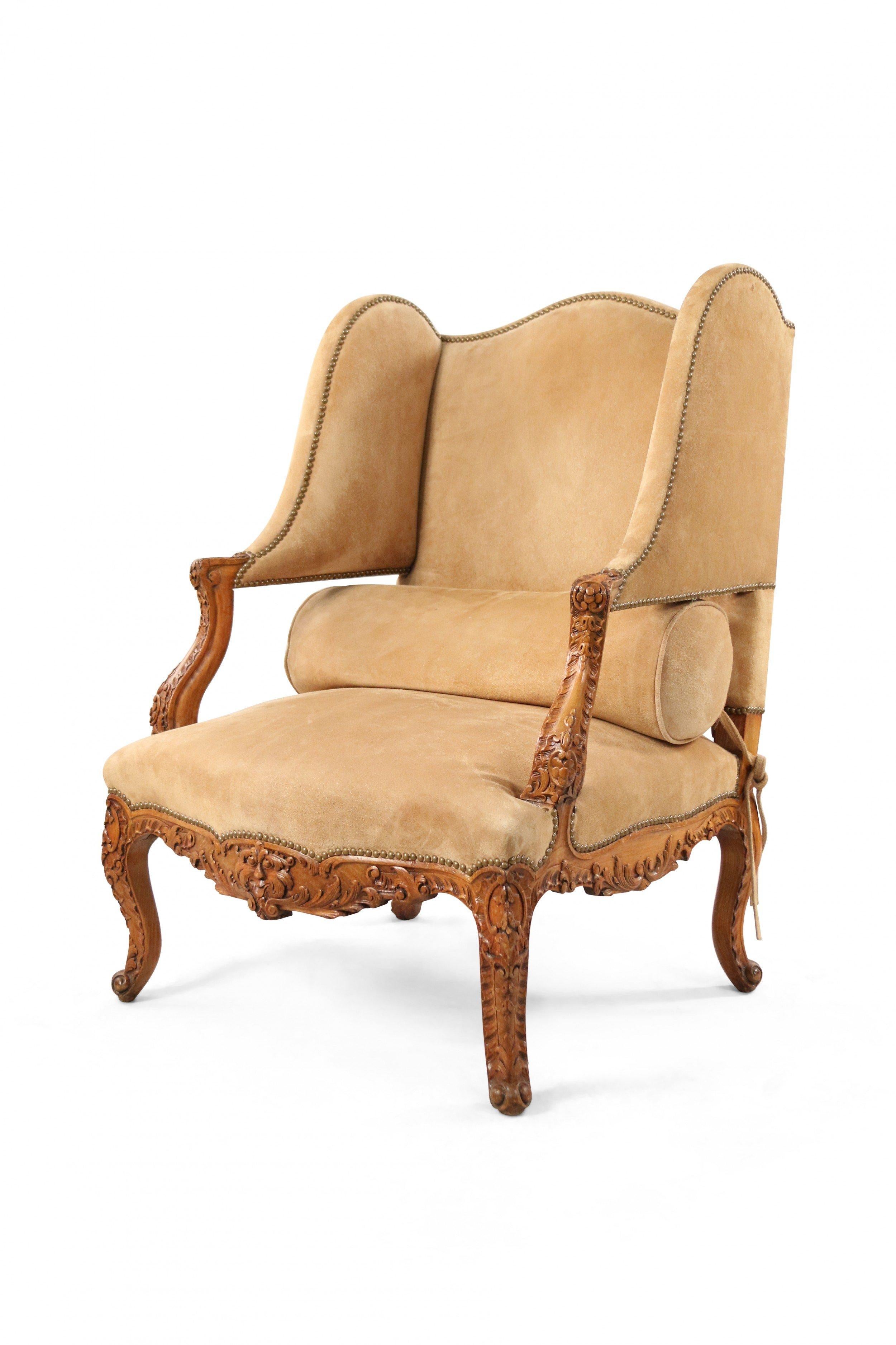 Pair of French Louis XV style (19th century) wing back open armchairs with an open lower back section beige suede upholstery, carved wooden arms and frames, and brass upholstery nail detail.