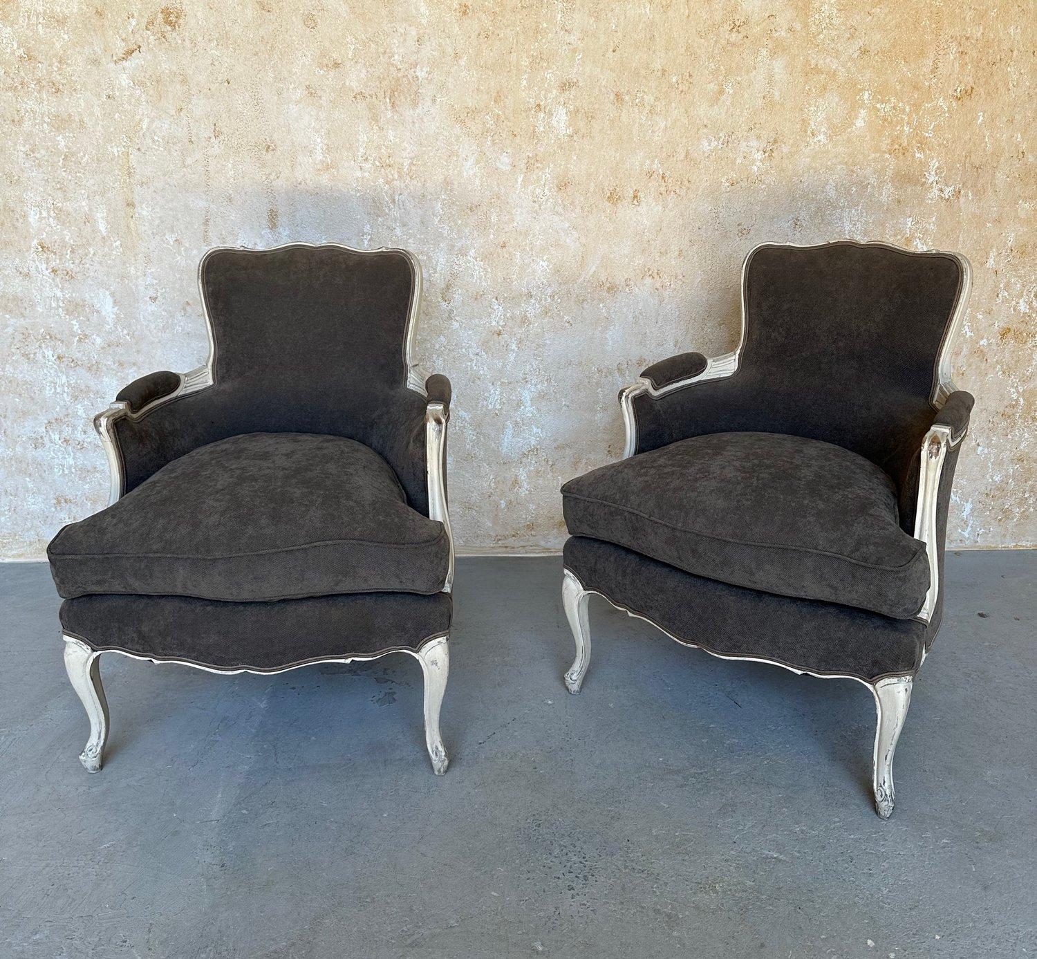 An elegant pair of French Louis XV style armchairs upholstered in gray fabric with matching piping. The ornate carved wooden frames are masterfully painted in a beautifully antiqued white patina that contrasts the rich gray upholstery. The padded