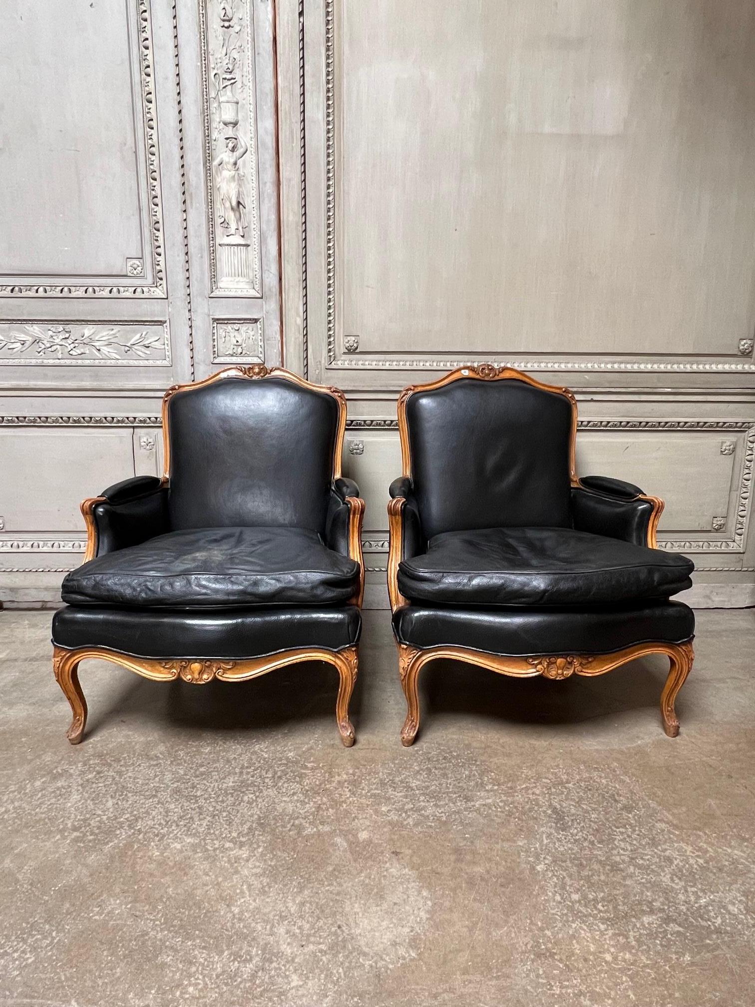 A large pair of French Louis XV style carved wood bergere armchairs with nice deep carving dating from the mid 20th century. These lounge chairs are currently covered in black leather but there is some wear on the seat cushions. they are usable but
