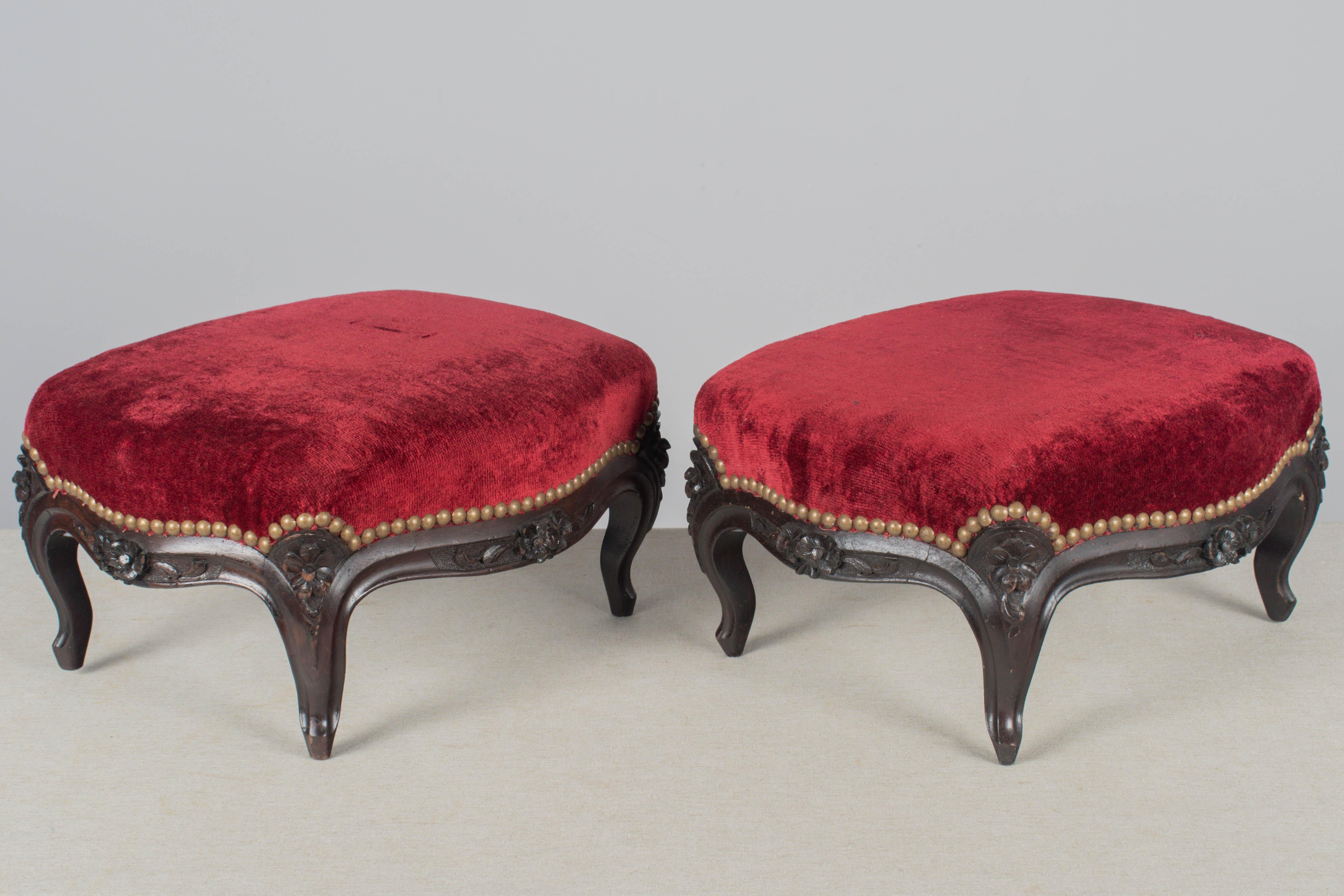 A pair of French Louis XV style upholstered foot stools with dark stained beech wood frames. Red velvet fabric is in good condition with some spotting. Brass nail head trim. Nice hand-carved floral details. Circa 1940-1950. 
Dimensions: 16