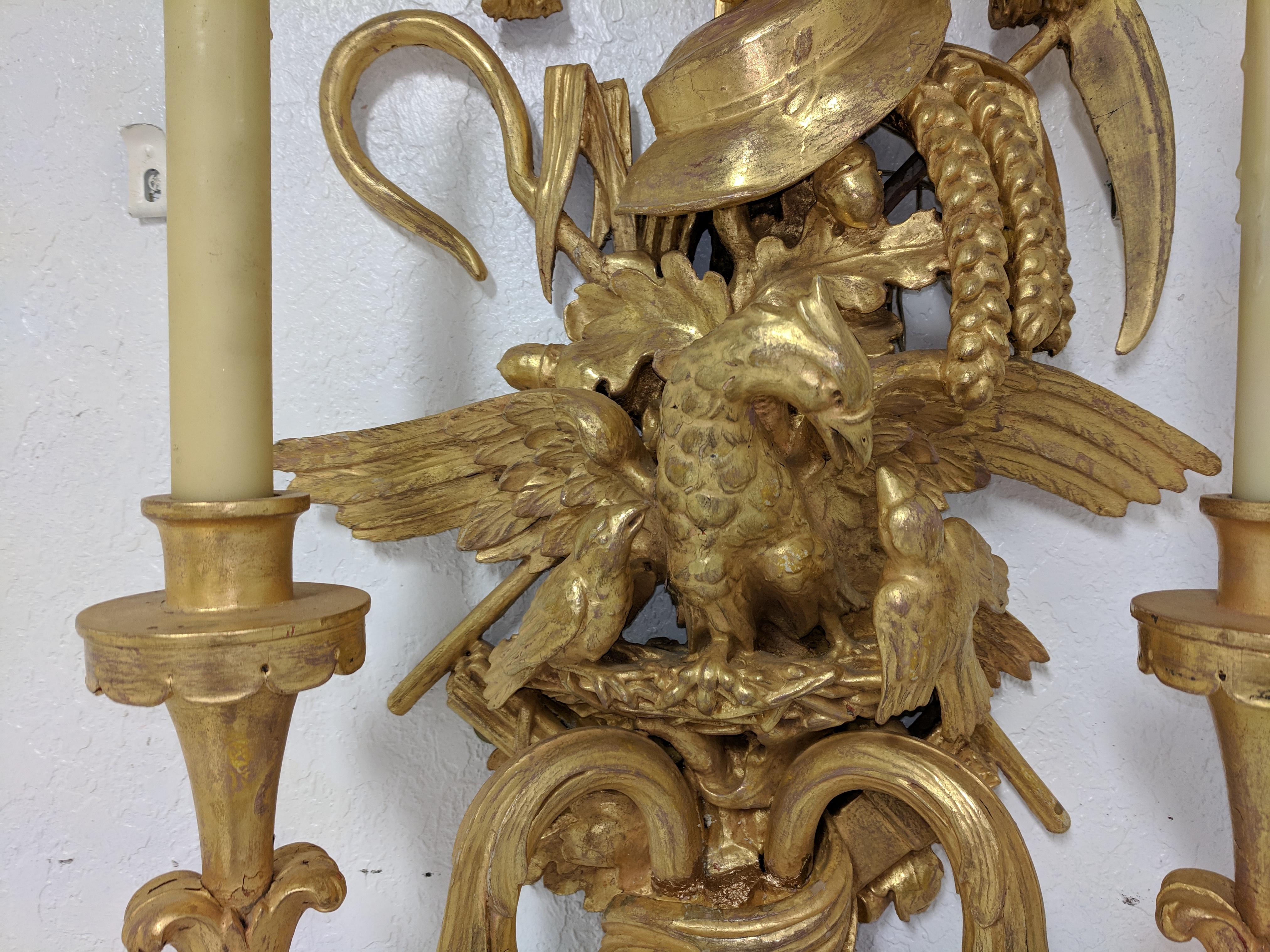 Tall pair of antique French Louis XV-style giltwood two-branch sconces.
Sconces feature a bow and two sets of tassels at top.
Beneath the tassels there is hat, a sythe, wheat sheaves, a hook,
corn husks and oak leaves with acorns.
A large bird